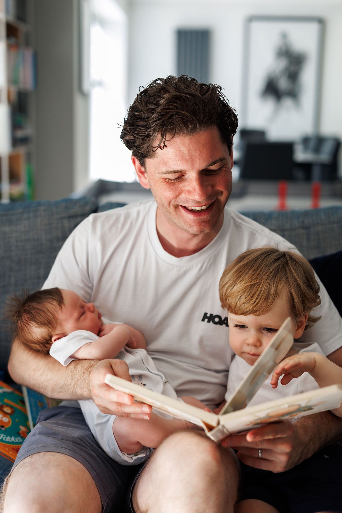 Smiling father reading book to toddler while holding sleeping newborn baby on couch at home.