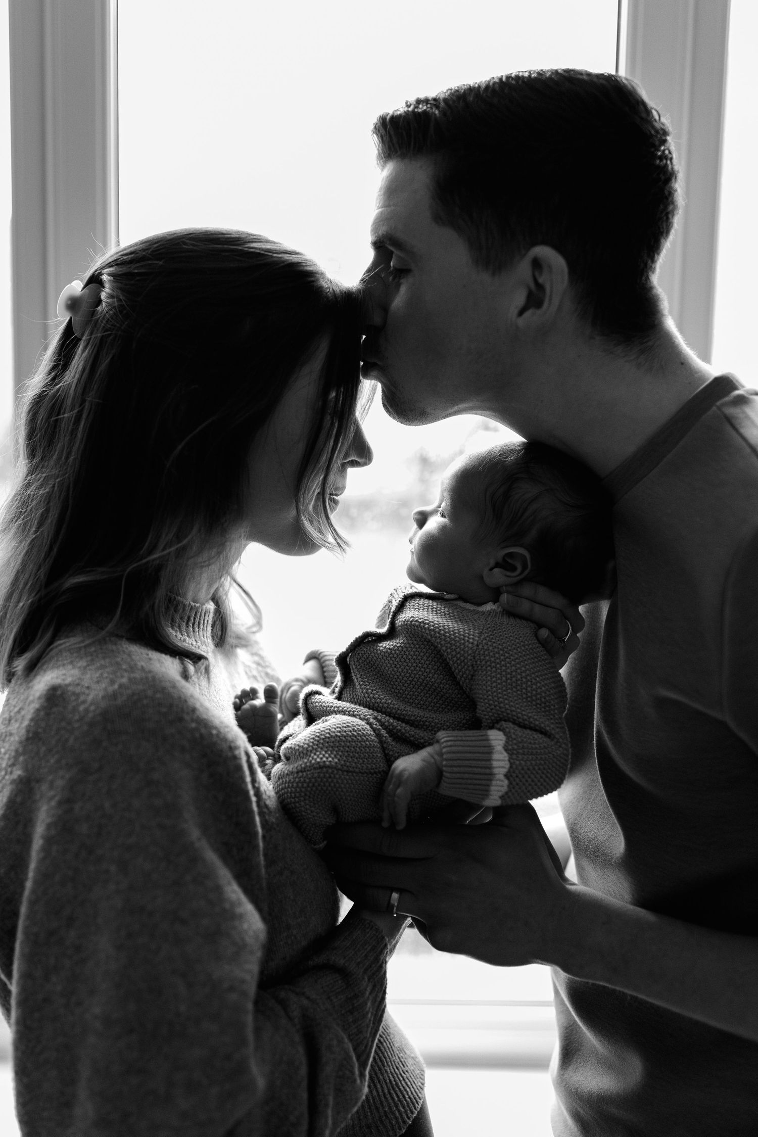 Black and white silhouette of new parents kissing while holding their newborn baby near a bright window.