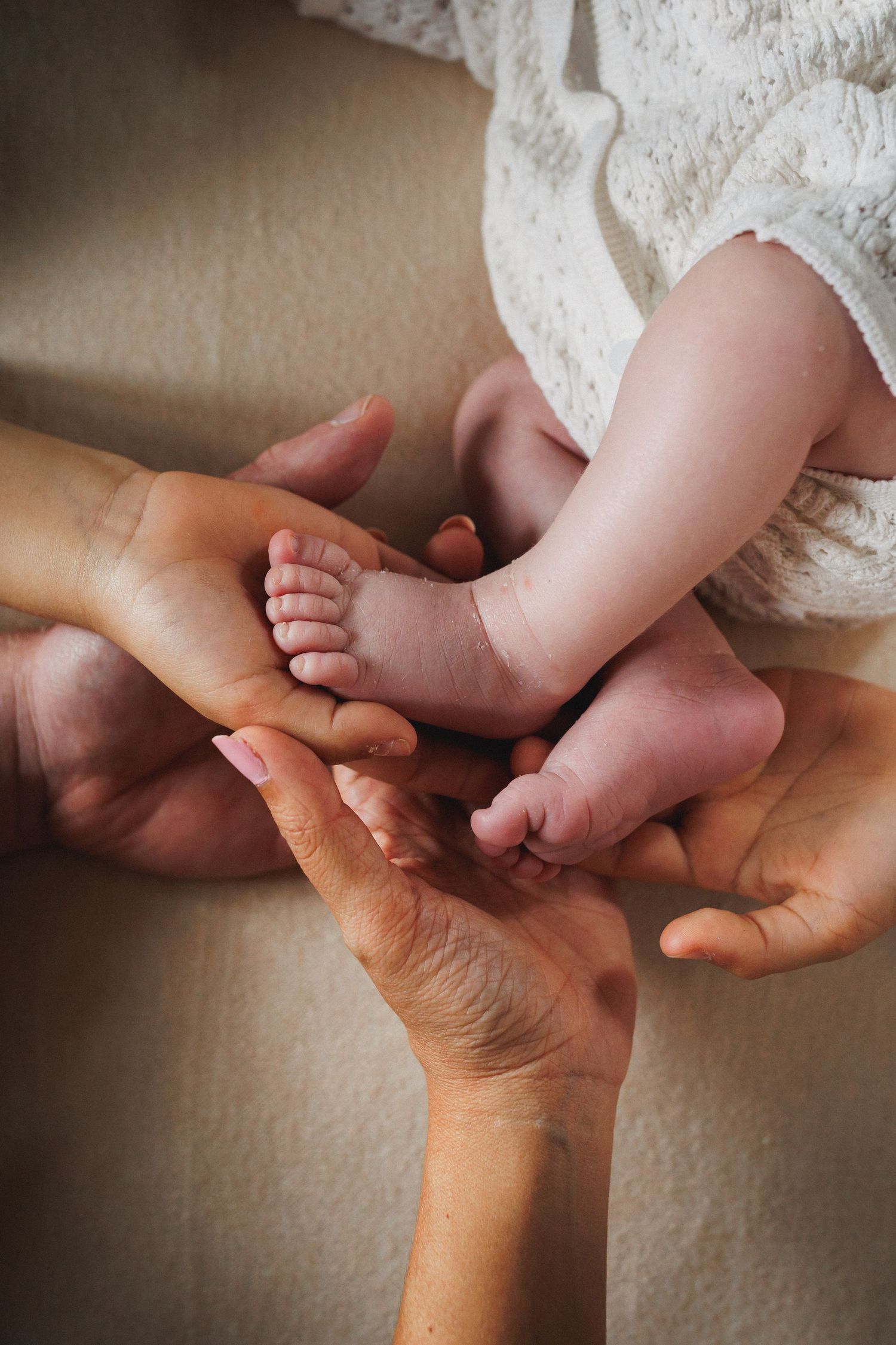 Two adult hands gently cradling newborn baby feet in soft white knit outfit.