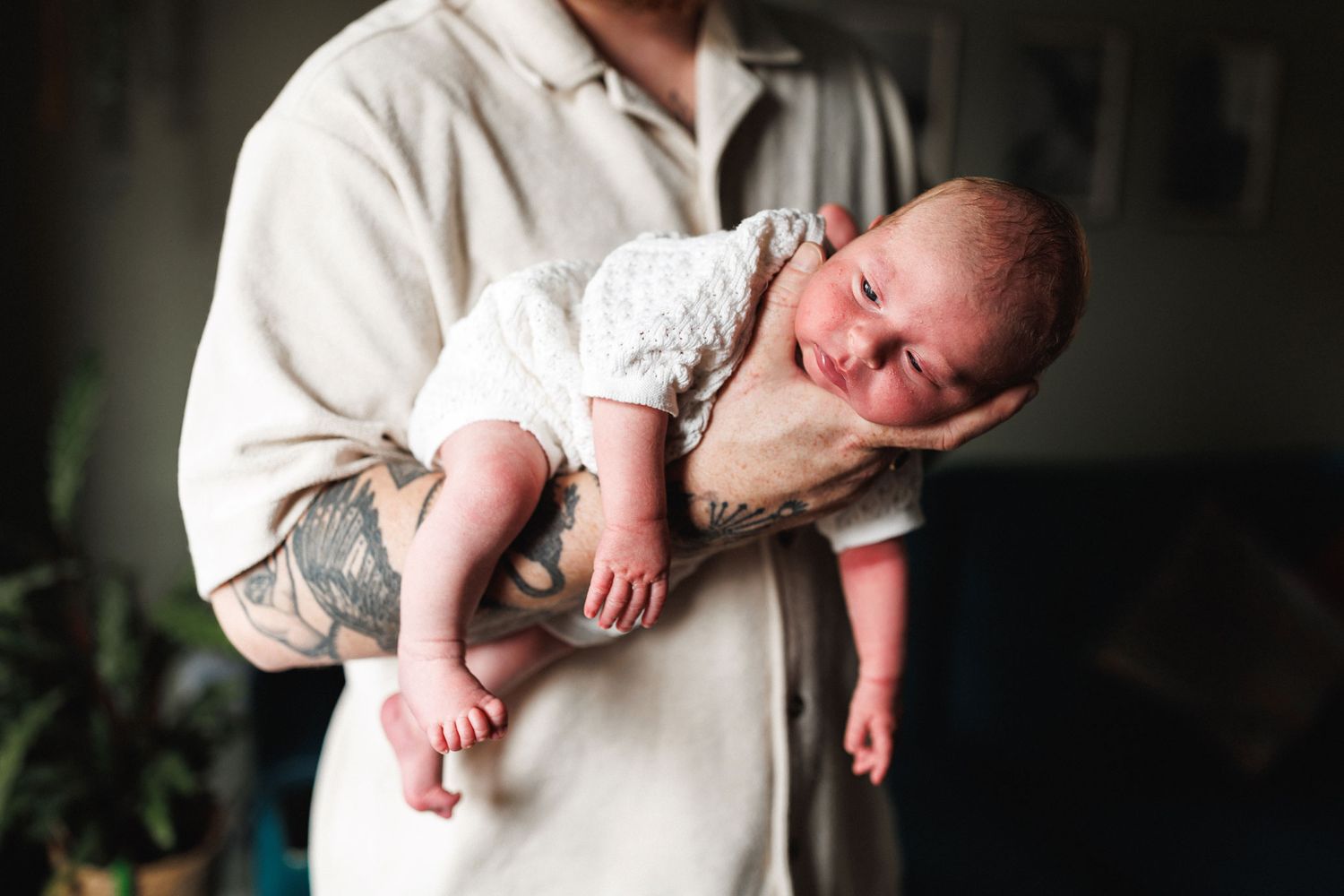 Tattooed parent cradling a sleepy newborn baby in their arms at home.
