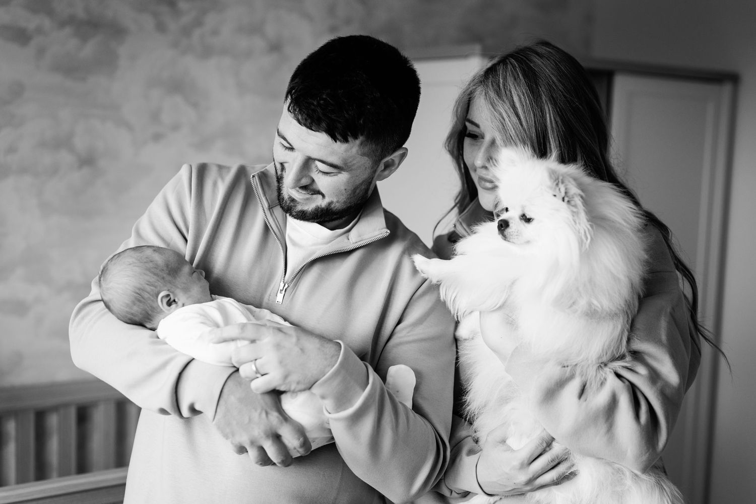 Black and white photo of a man holding a newborn baby while a woman holds a white Pomeranian dog.