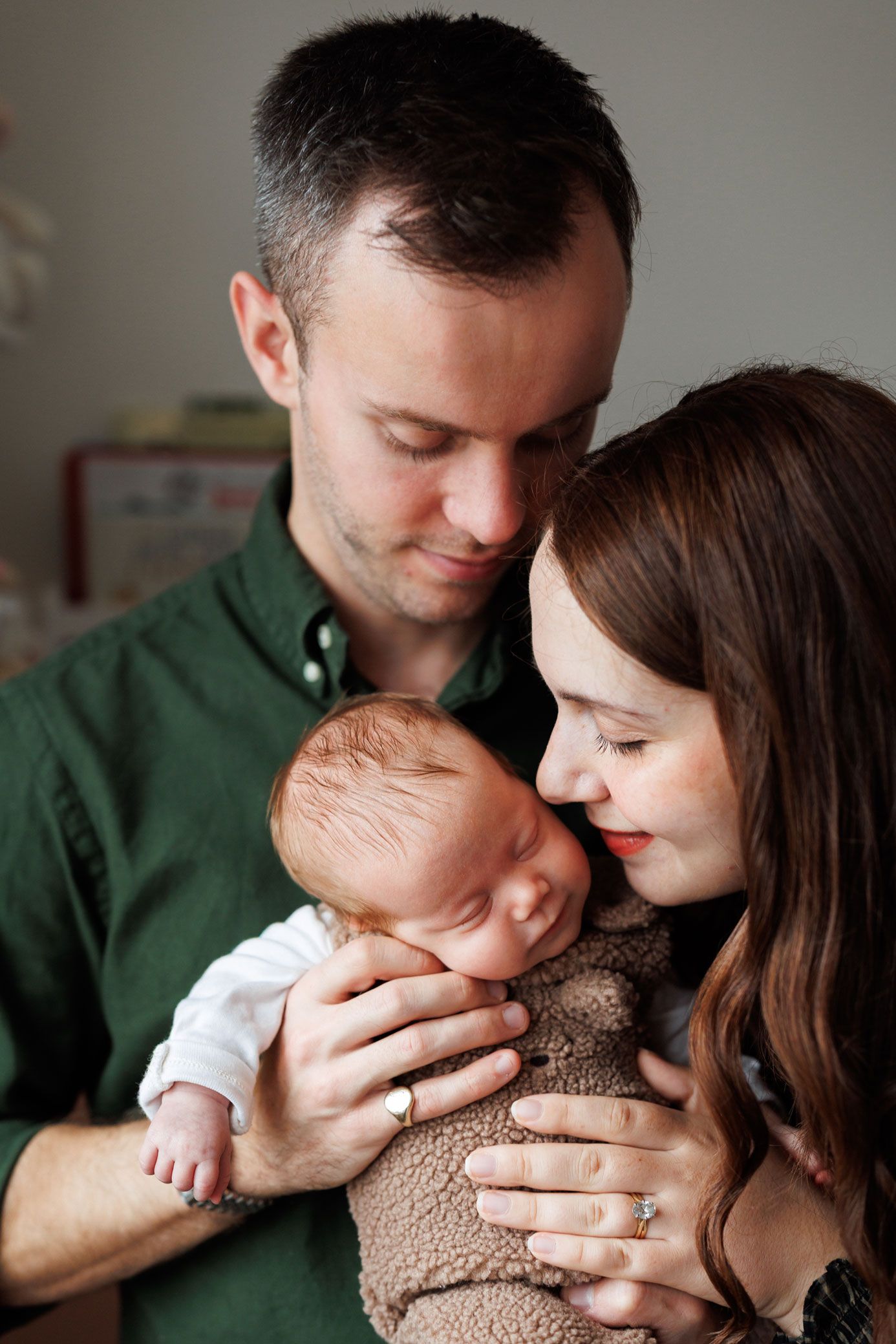 A smiling mother and father in a green shirt gently cradle their sleeping newborn baby wrapped in a soft brown knit blanket.