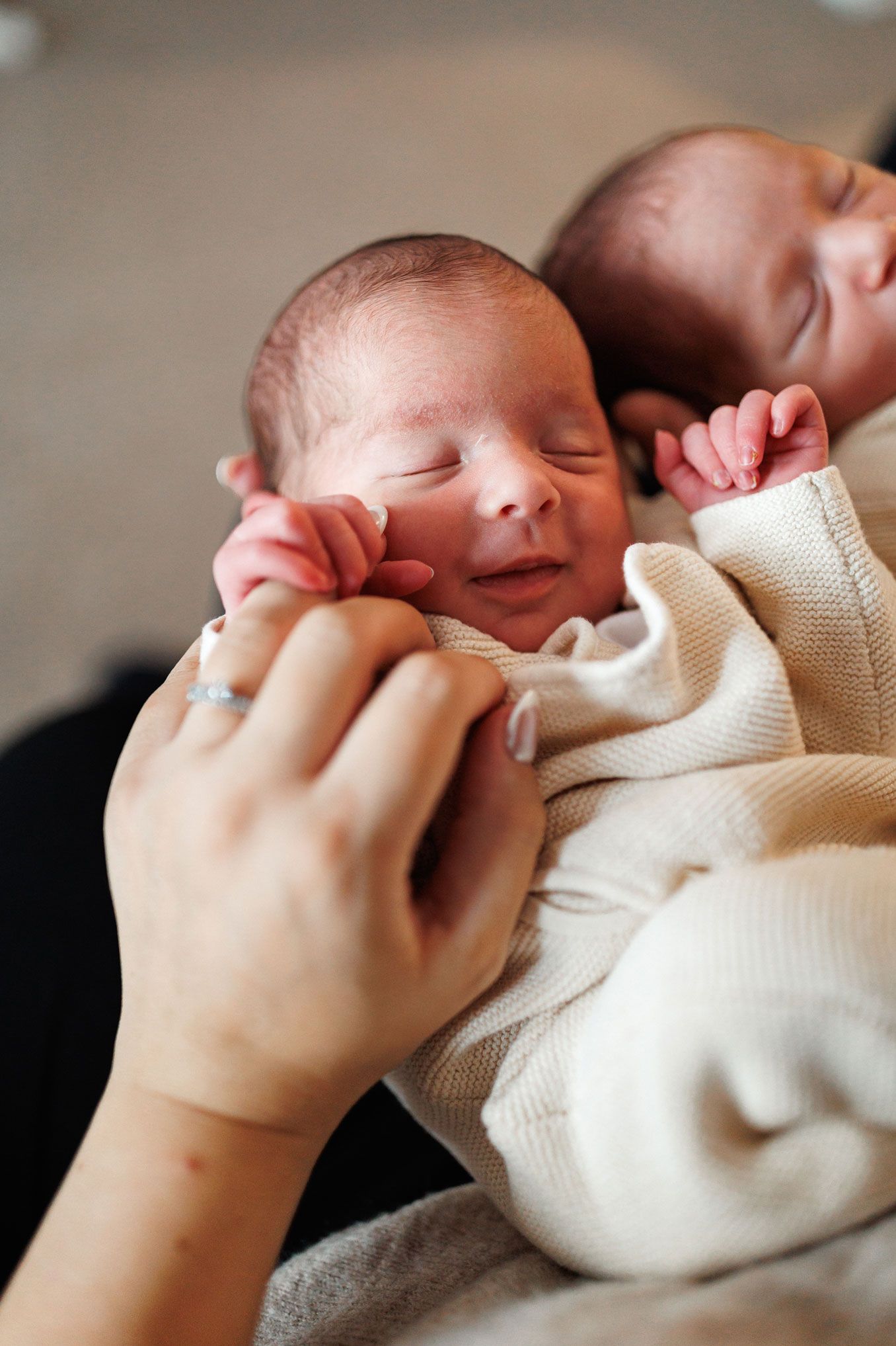 Smiling newborn twin in a cream knit outfit gently held by a hand with a silver ring, with a sleeping sibling beside them.