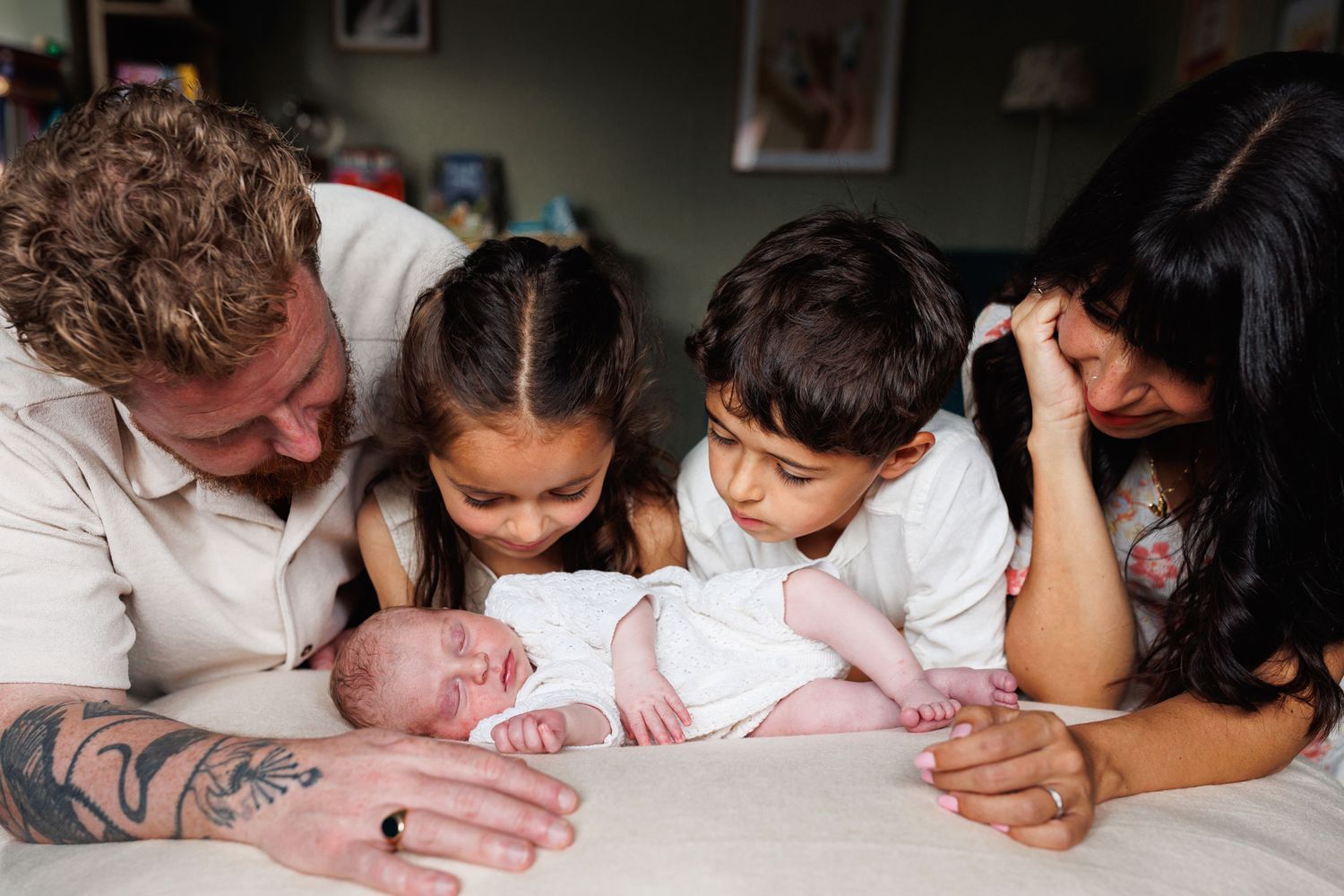 Family of four with tattoed father, mother, and two children admiring their newborn baby on a bed.