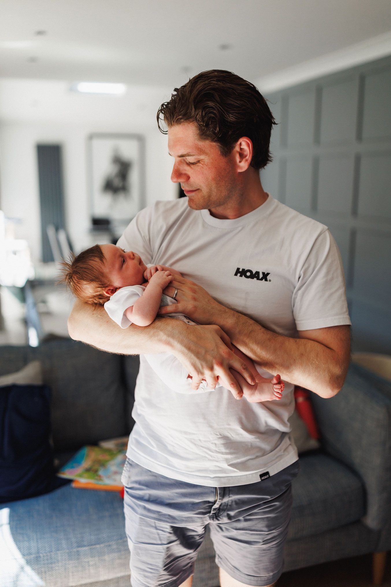 A father in a white HOAX t-shirt tenderly holds his newborn baby in a modern living room.