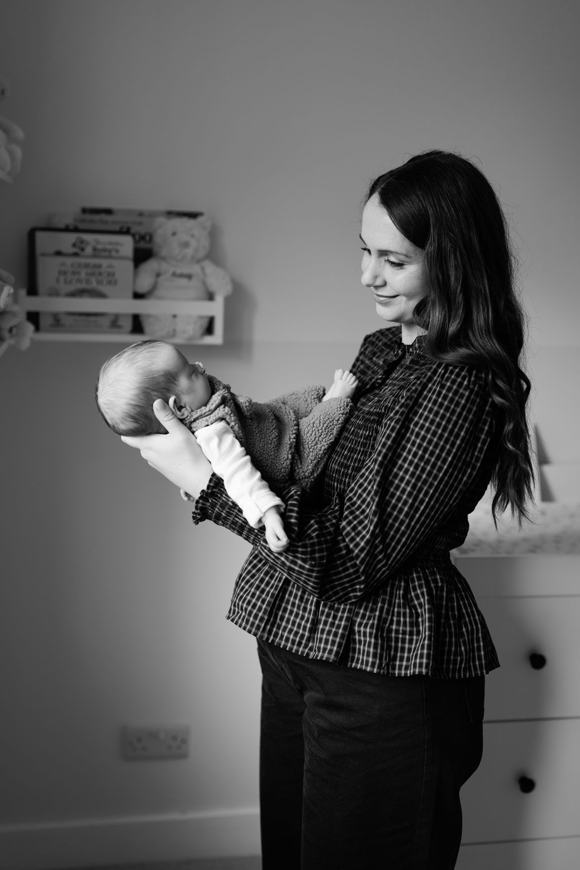 Black and white photo of a smiling mother in a plaid blouse cradling her newborn baby in a nursery with a teddy bear on a shelf.