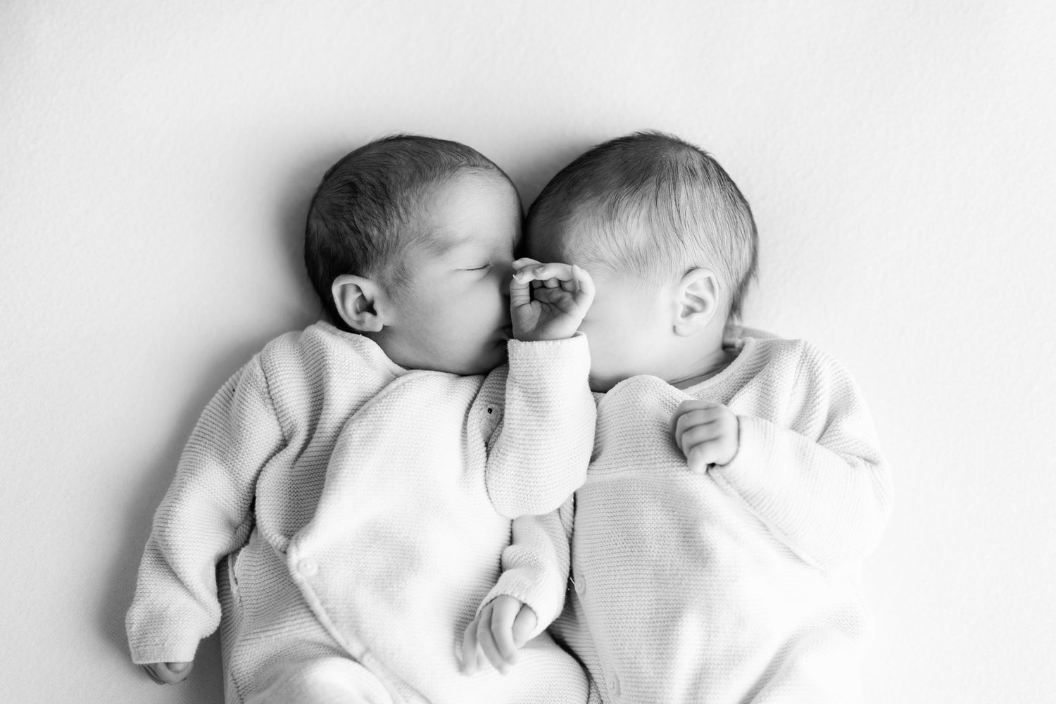 Black and white photo of two sleeping newborn twins in soft knit outfits lying side by side, their tiny hands touching.
