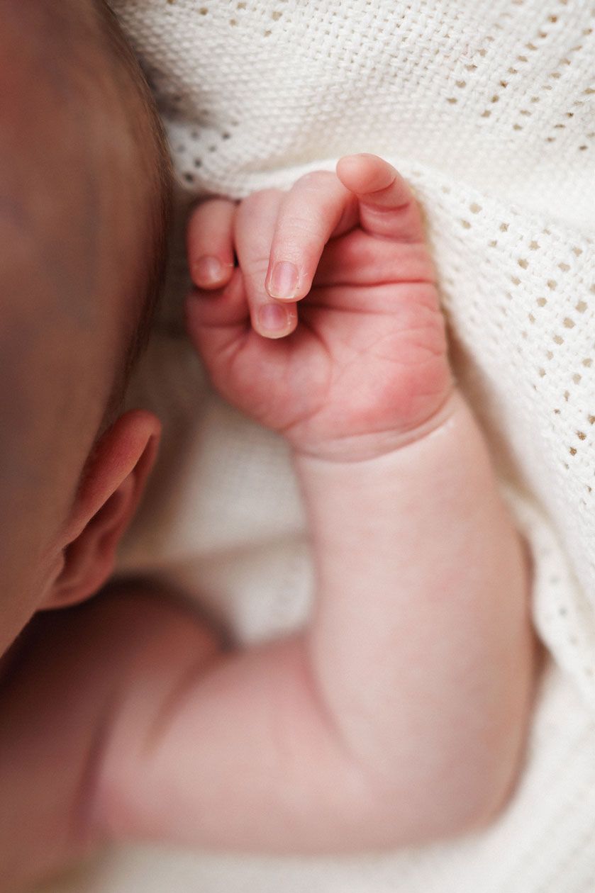 Close-up of a sleeping newborn baby's tiny clenched fist resting against a soft white knit blanket.
