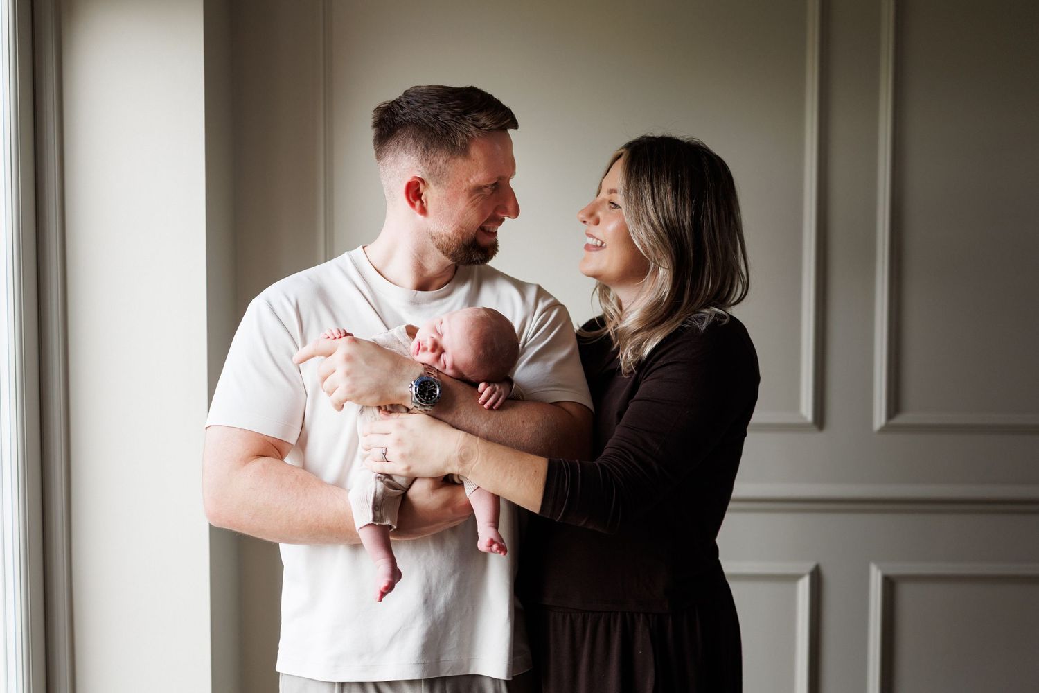 A smiling couple stands in a bright room with paneled walls, gently holding their sleeping newborn baby between them.