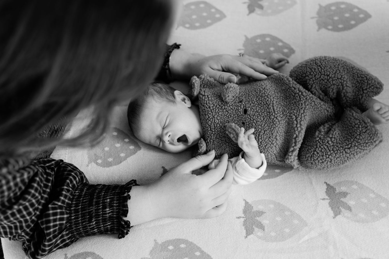 Black and white photo of a mother gently touching a sleeping newborn baby in a fuzzy bear onesie on a strawberry-print blanket.