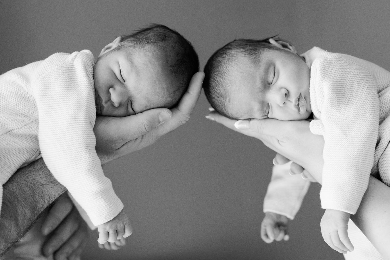 Two sleeping newborn babies in white outfits rest their heads on adult hands in a soft black and white photo.