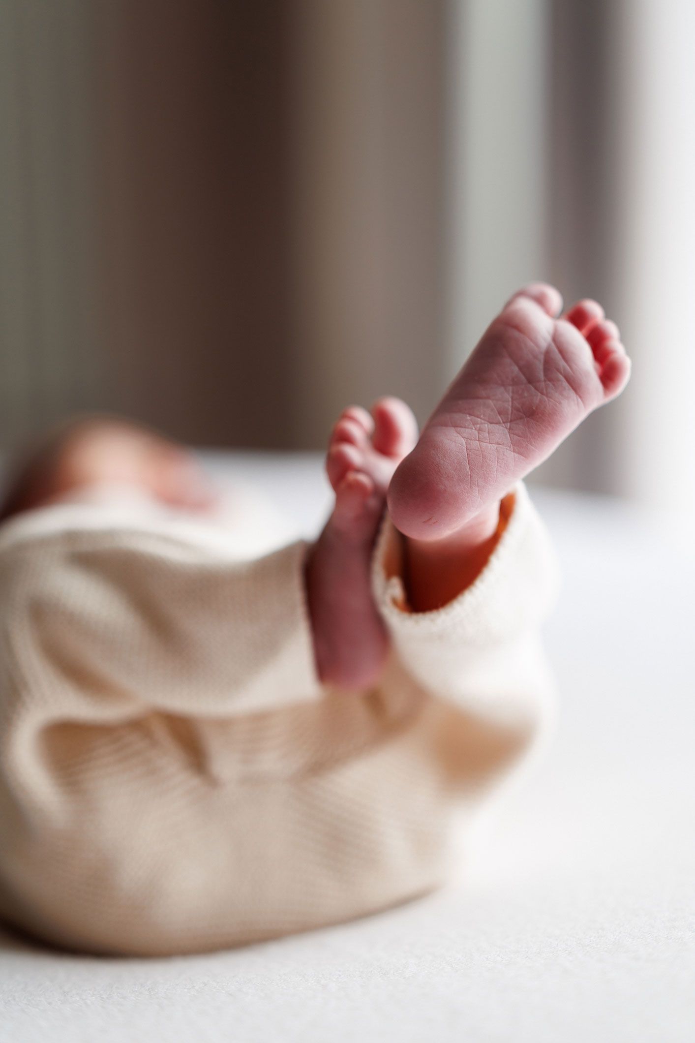Close-up of a newborn baby's tiny bare feet in focus, wrapped in a soft cream knit outfit and resting on a white blanket.