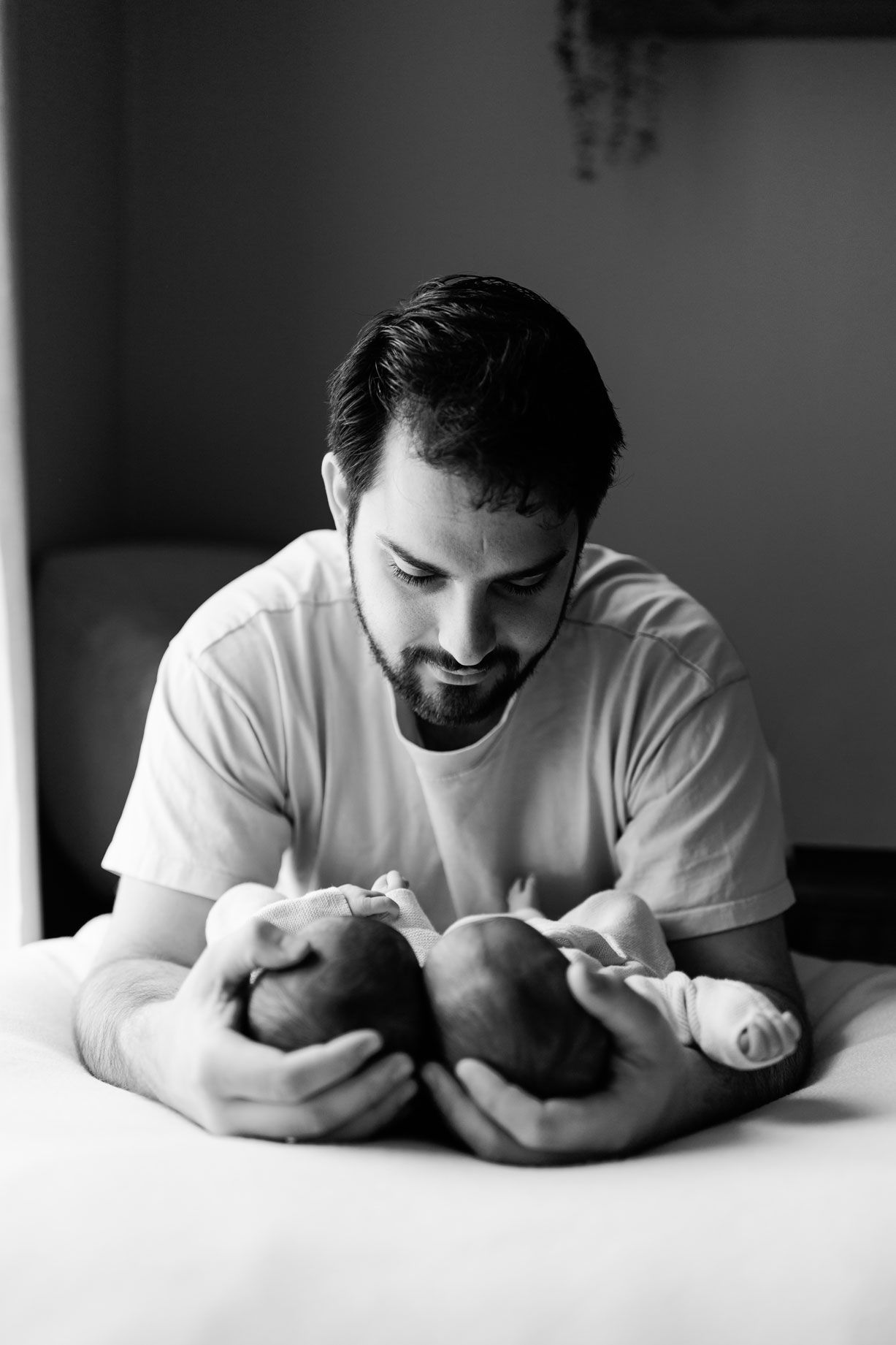 Black and white photo of a bearded man in a t-shirt gently cradling two sleeping newborn babies on a bed.