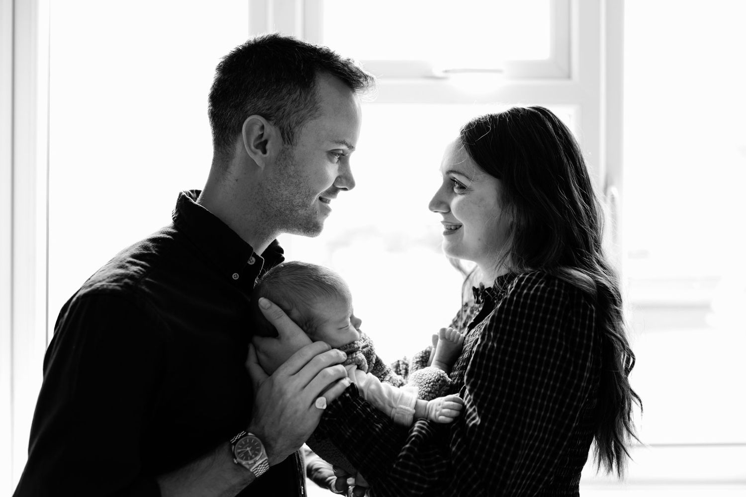 Black and white photo of smiling parents gazing at each other while cradling their sleeping newborn baby beside a bright window.