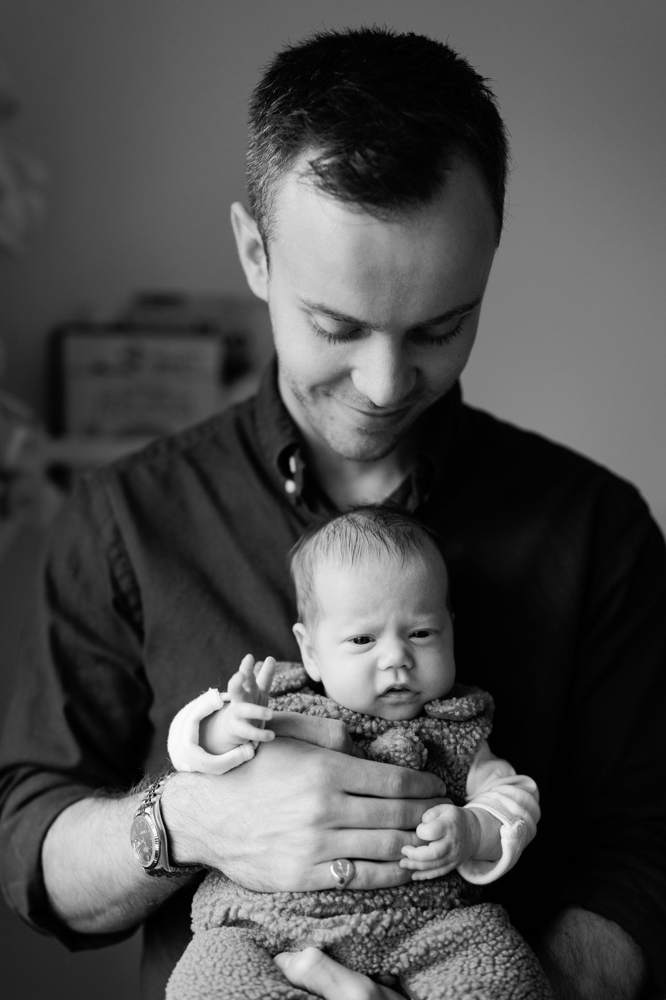 Smiling man in a dark button-up shirt gently cradles a wide-eyed newborn baby in a cozy knit outfit in a black and white portrait.