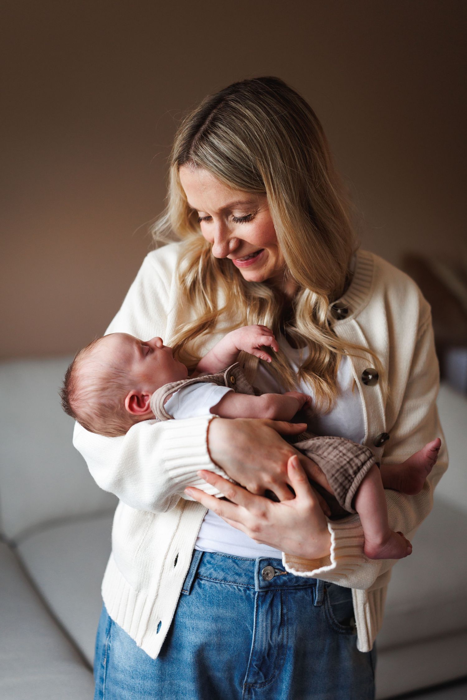 Smiling mother in white cardigan tenderly holding her newborn baby in a cozy home setting.