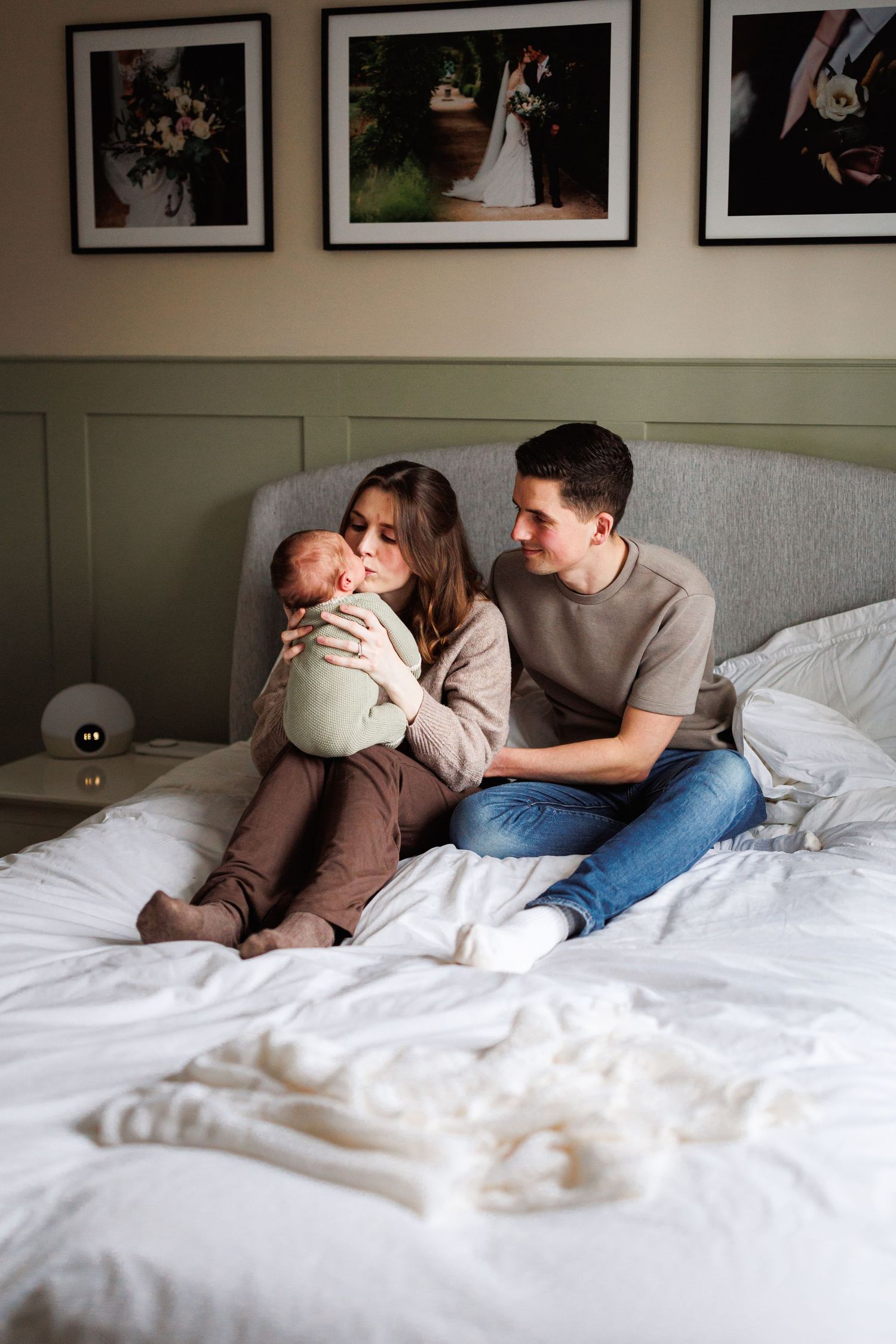 A couple sits on a white bed as the mother kisses their newborn baby while the father watches lovingly.