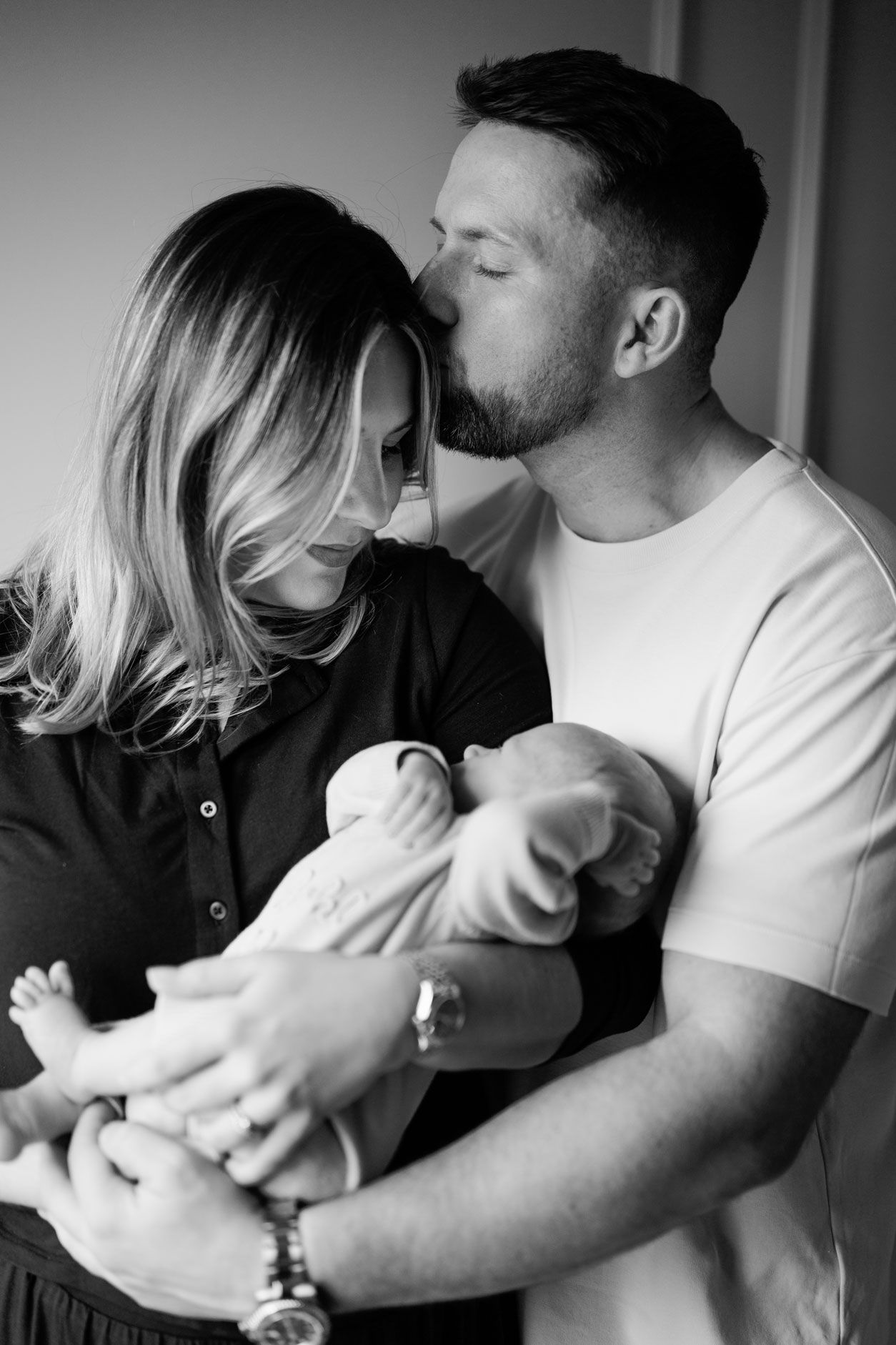 Black and white portrait of a father kissing his partner's forehead as she gently cradles their sleeping newborn baby in her arms.
