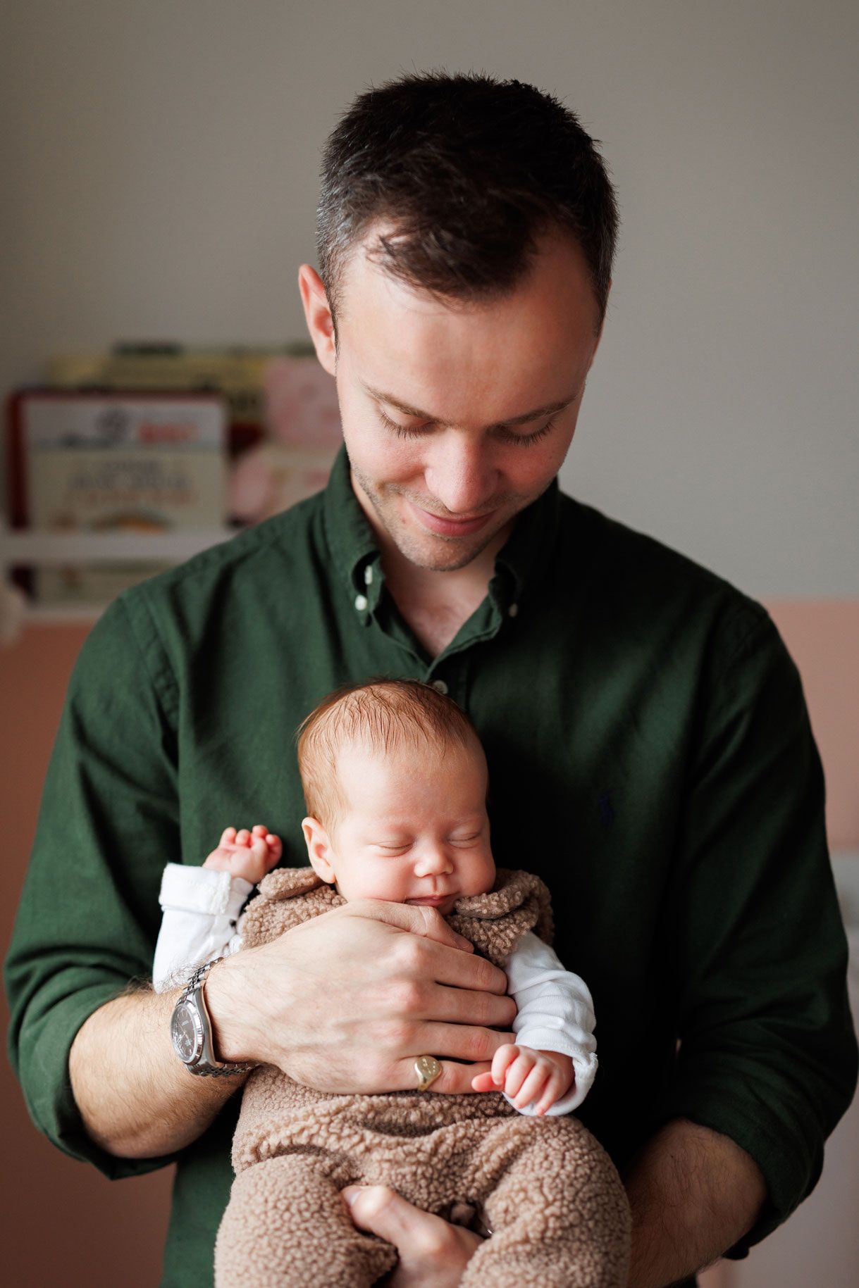 A smiling man in a green button-up shirt gently cradles a sleeping newborn baby dressed in a soft brown outfit.