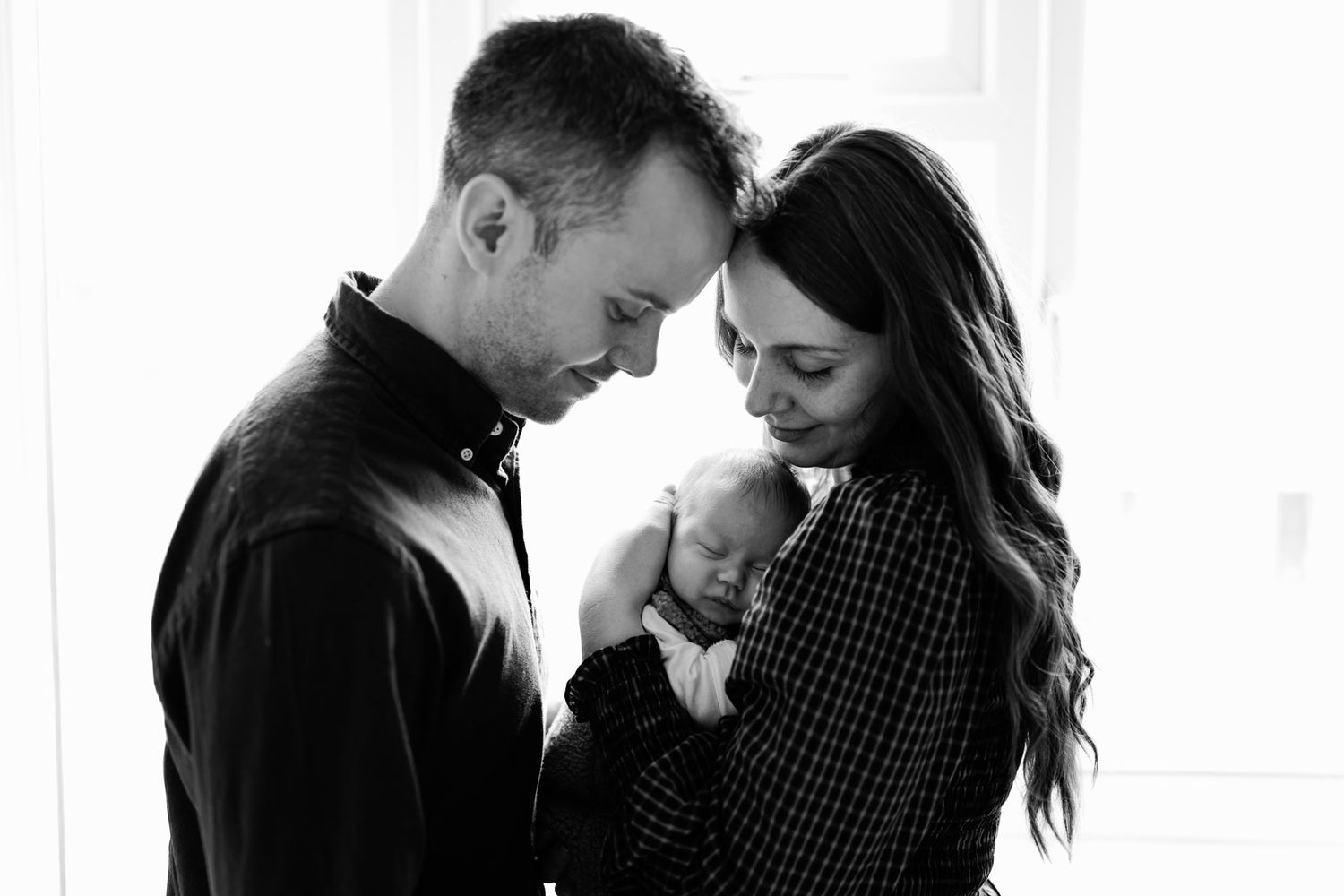 Black and white newborn photo of parents standing foreheads together, gently cradling their sleeping baby in a bright, softly lit room.