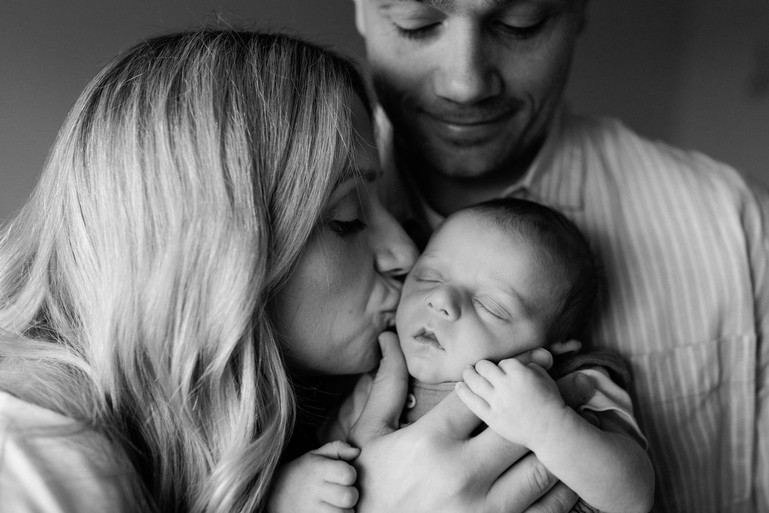 Black and white photo of parents lovingly kissing their newborn baby together.