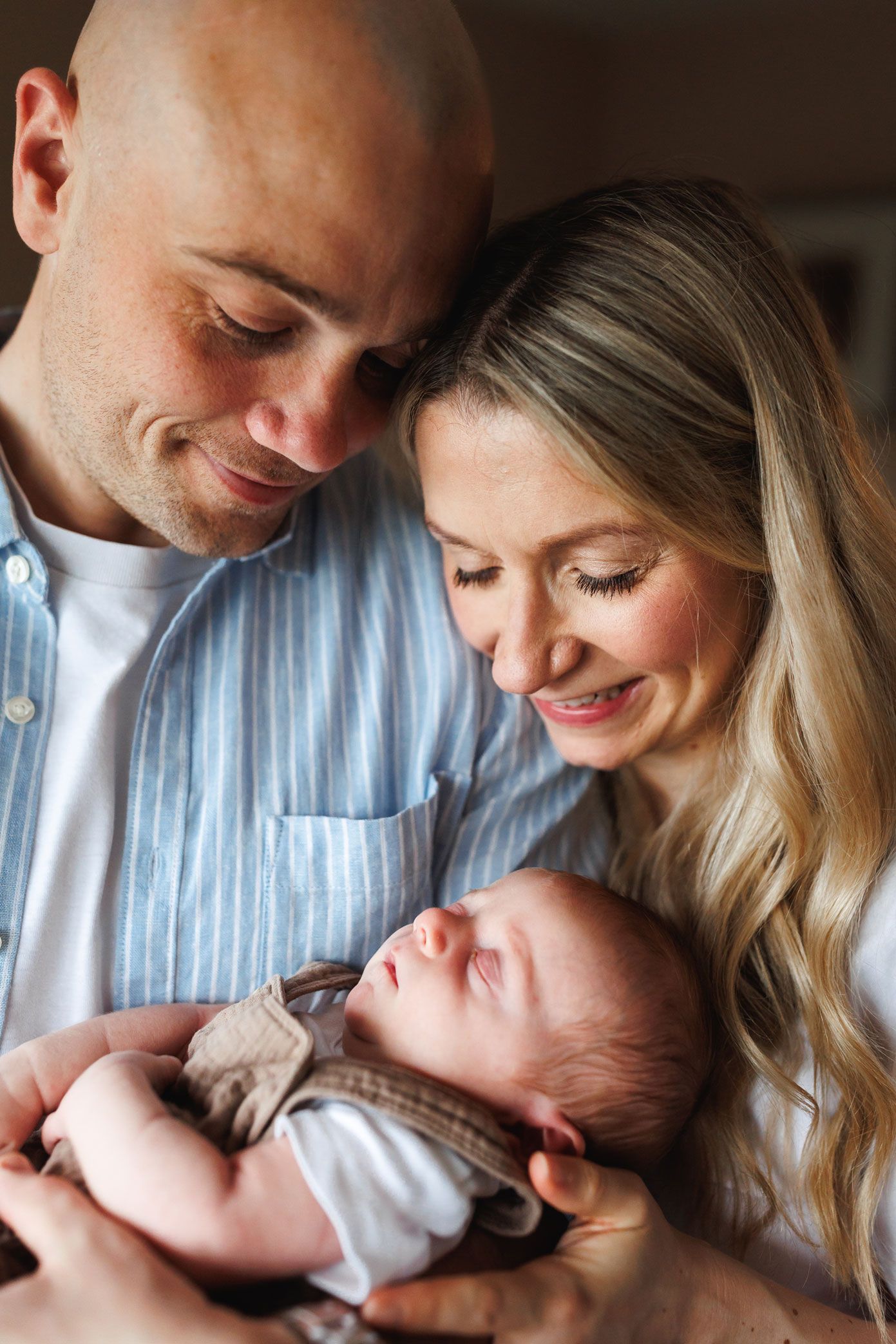 Happy parents smiling at their newborn baby, with father in blue striped shirt and mother with blonde hair.