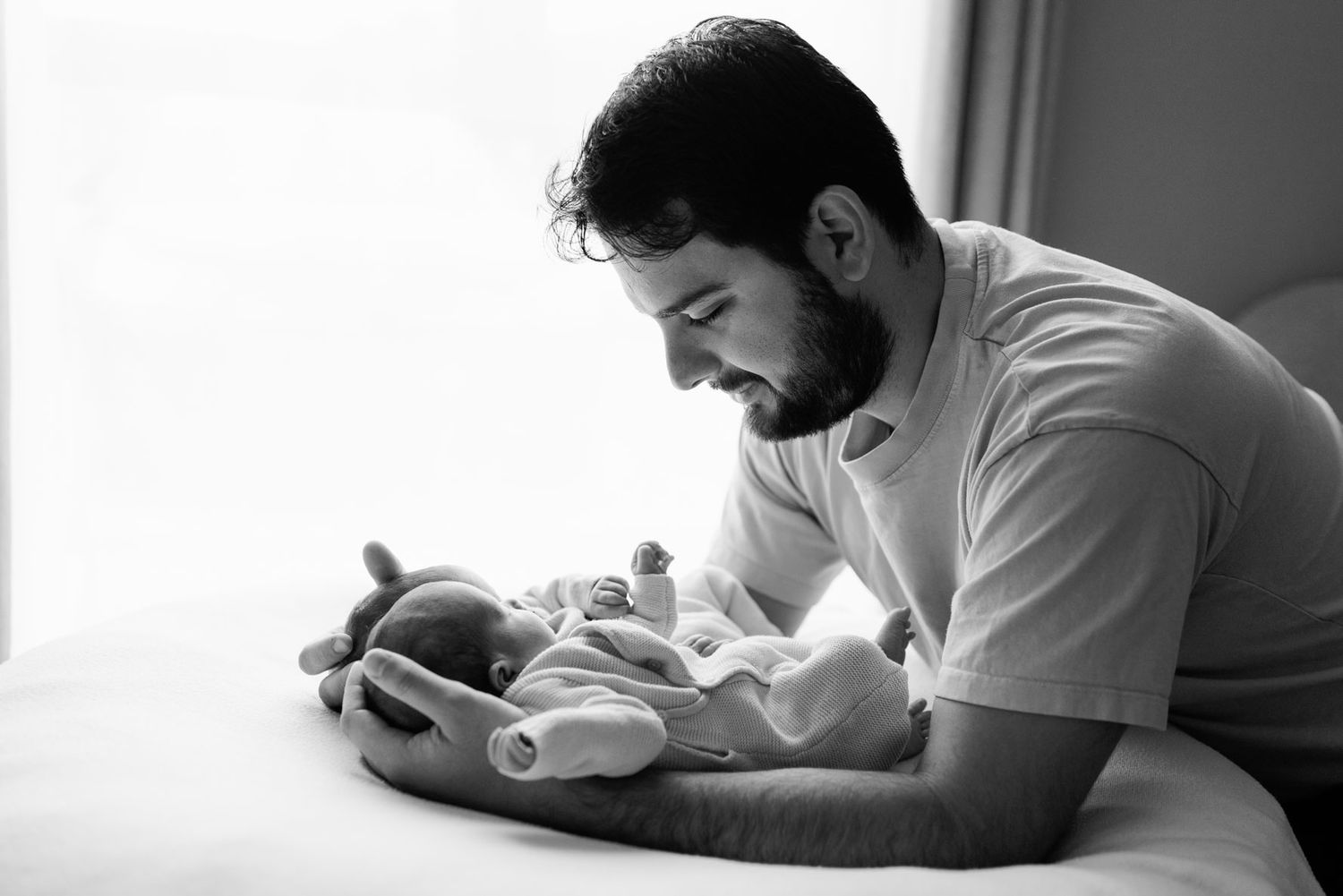Black and white photo of a bearded father gazing lovingly at his sleeping newborn baby wrapped in a blanket on a bed by a bright window.