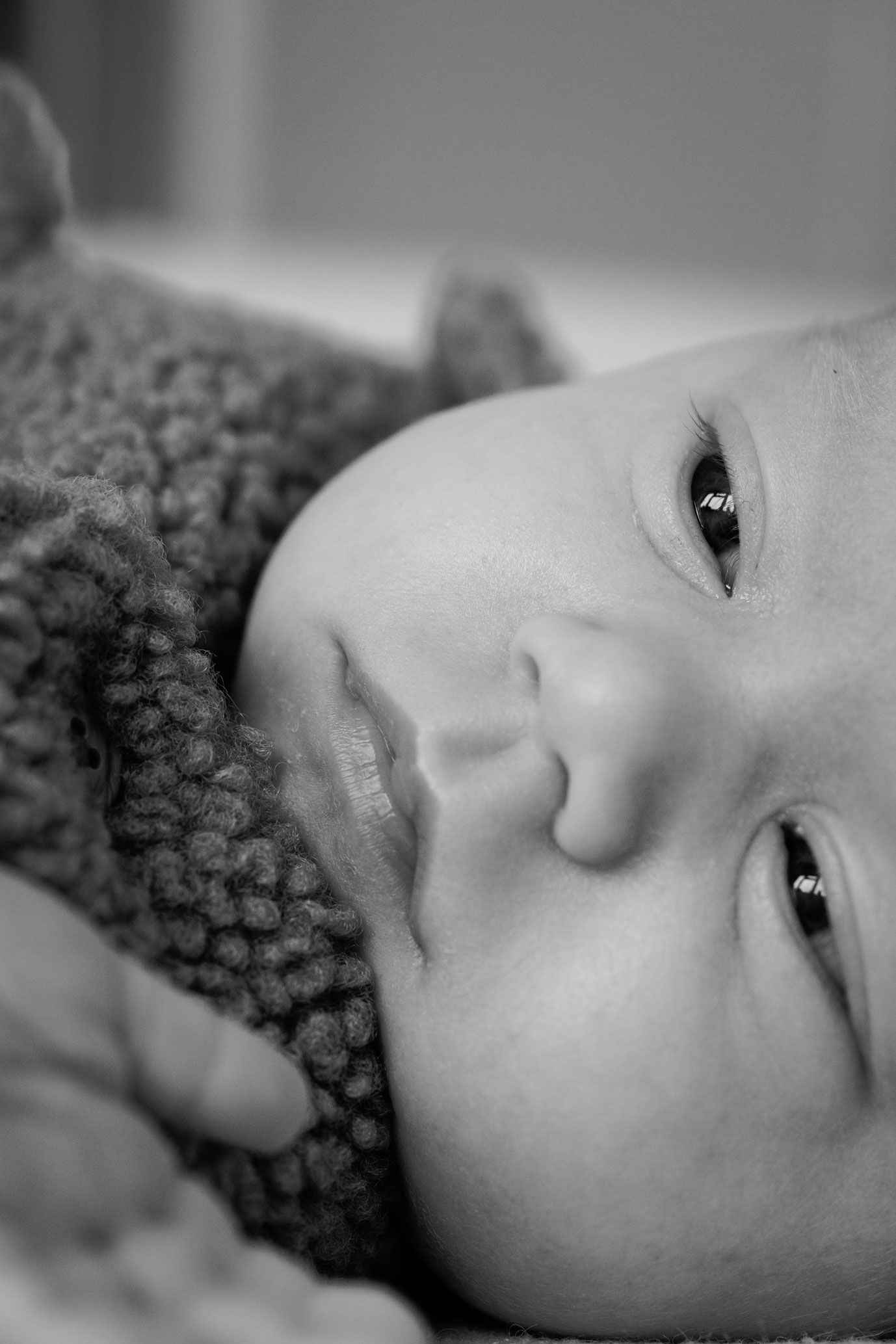 Black and white close-up portrait of a newborn baby with wide eyes lying on a soft knitted blanket.