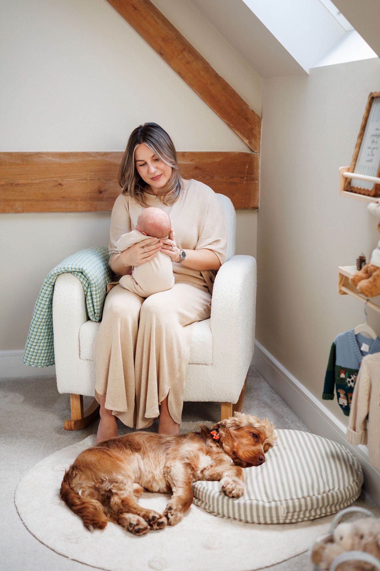 Woman in a beige outfit holds a sleeping newborn in a cozy armchair while a cocker spaniel naps on a rug in a bright nursery.