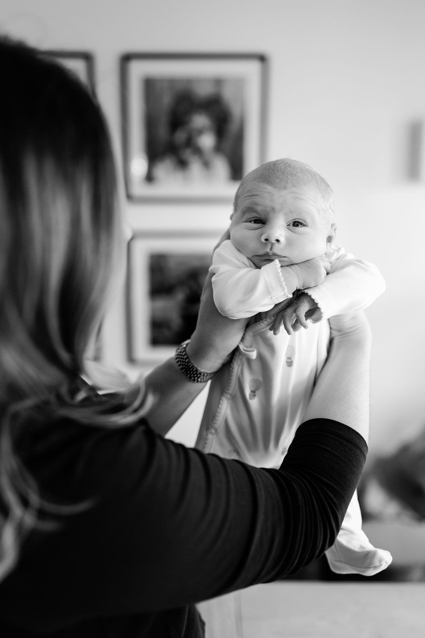 Black and white photo of a woman with long hair holding up a wide-eyed newborn baby in a onesie near framed pictures on the wall.