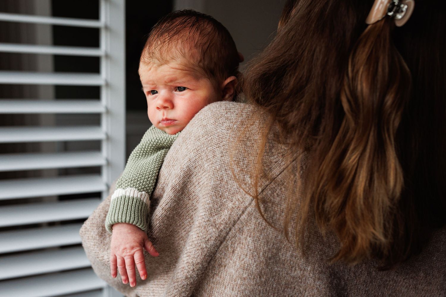 A mother in a beige knit sweater gently holds her newborn baby dressed in a green outfit near a window.