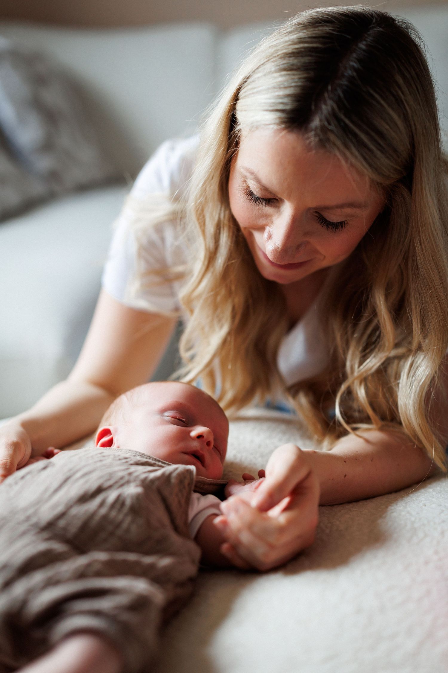 A smiling blonde mother gazes lovingly at her sleeping newborn baby on a white blanket.