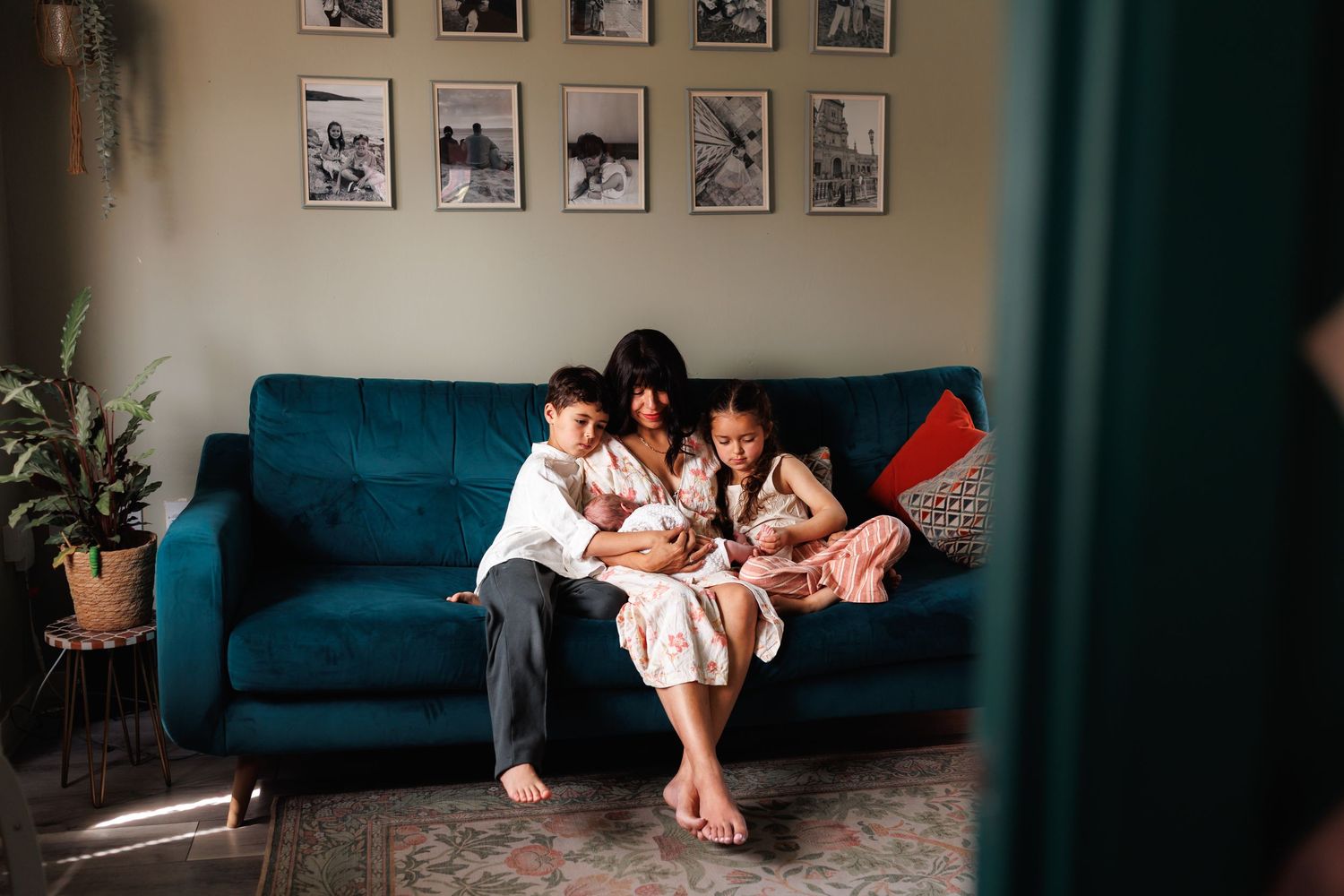A mother sits on a teal sofa with her two children, tenderly holding a newborn baby in their cozy home.