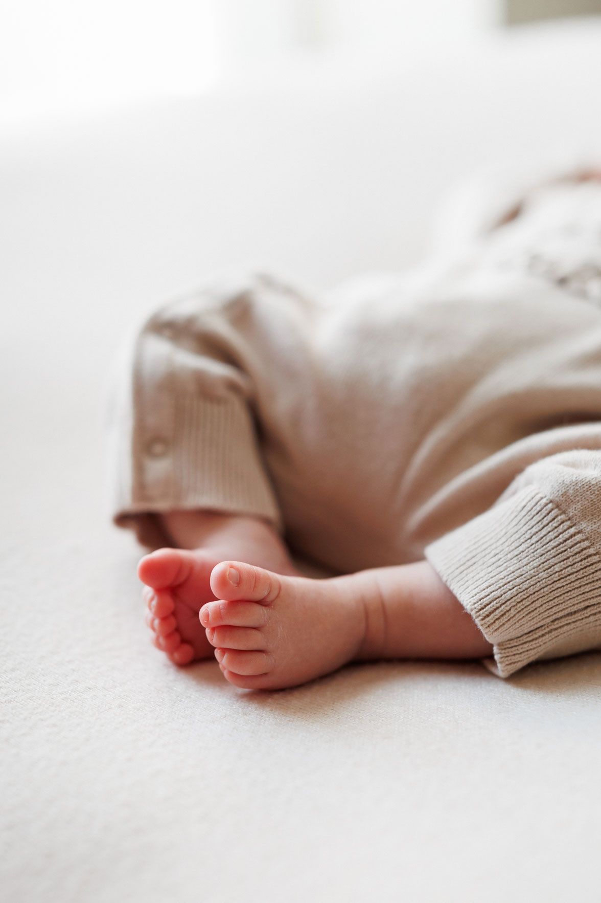 Close-up of a newborn baby's tiny bare feet resting on a soft cream blanket, dressed in a cozy beige knit outfit.