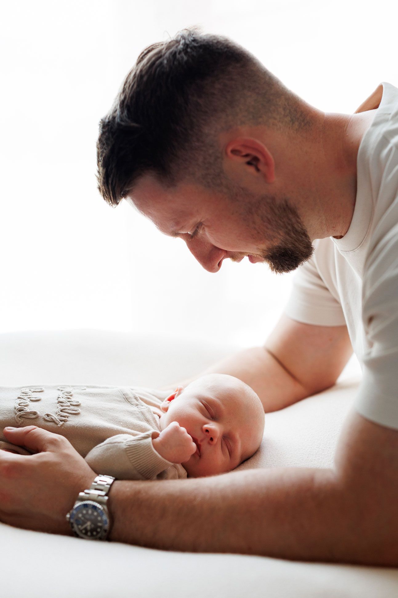 A father with short dark hair and a beard gazes lovingly down at his sleeping newborn baby resting on a soft white blanket.