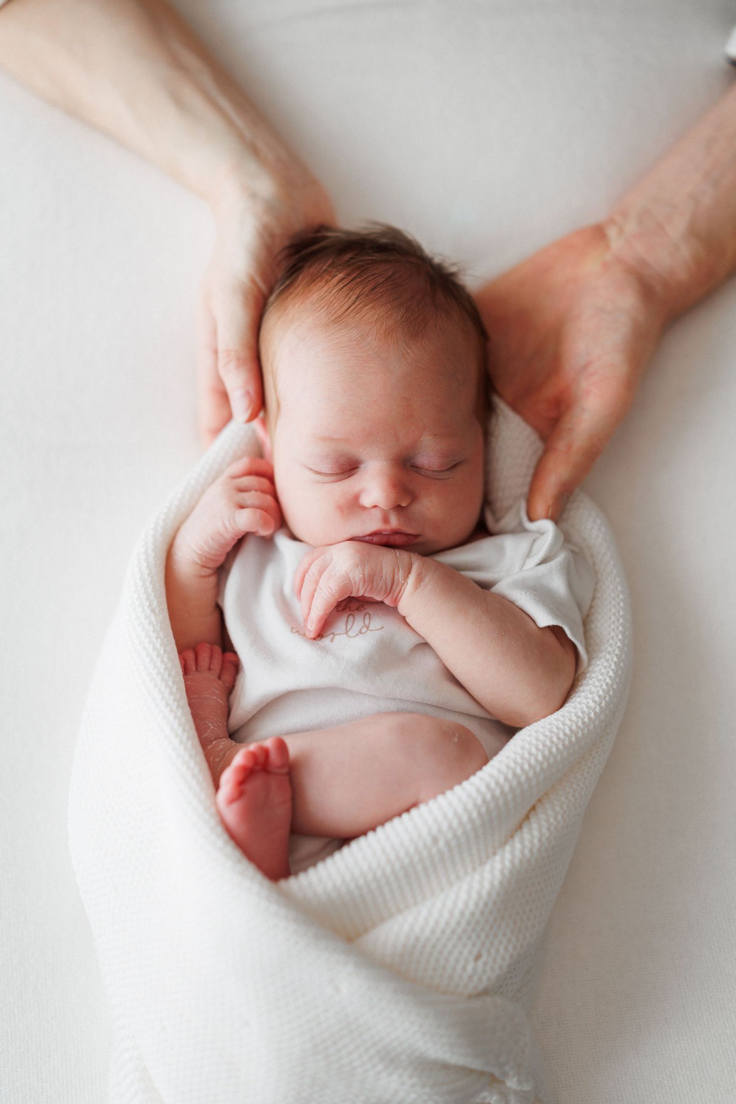 Sleeping newborn baby wrapped in a soft white knit blanket while two parent hands gently cradle the baby's head.