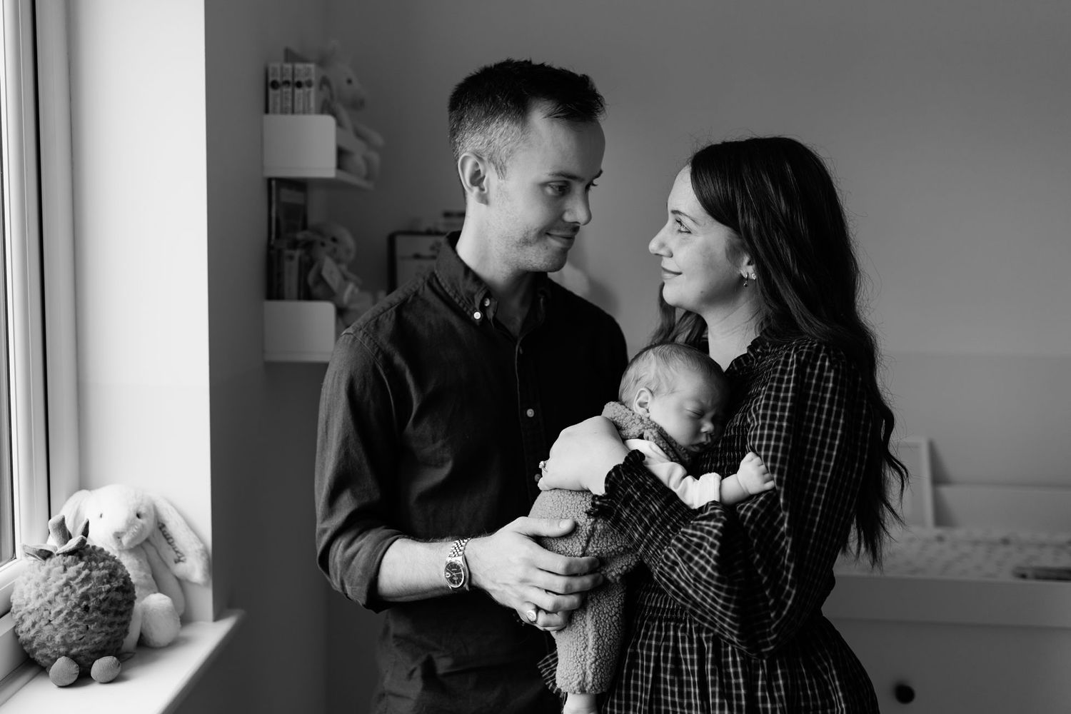 Black and white photo of smiling parents gazing at each other while holding their newborn baby in a cozy nursery beside a sunlit window.