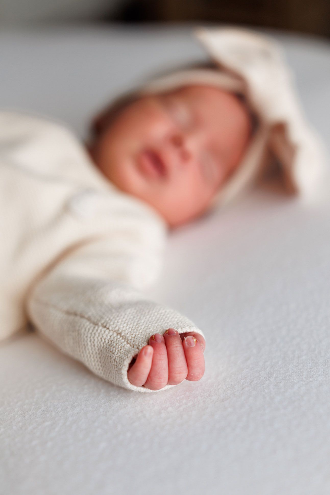 Close-up of a newborn's tiny hand peeking from a cream knit sleeve, with the baby sleeping softly out of focus behind.