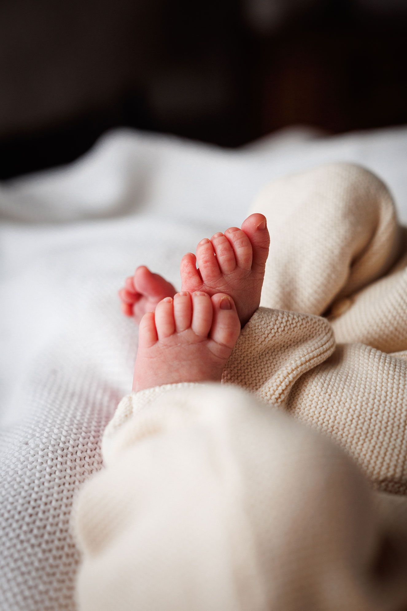 Tiny newborn baby feet with delicate toes peeking out from a cream knit outfit on a soft white woven blanket.