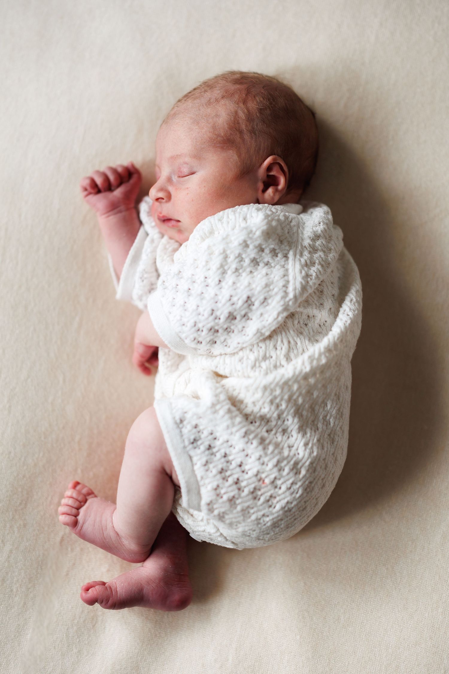 A peacefully sleeping newborn baby dressed in a white knit outfit lies on a soft cream blanket.