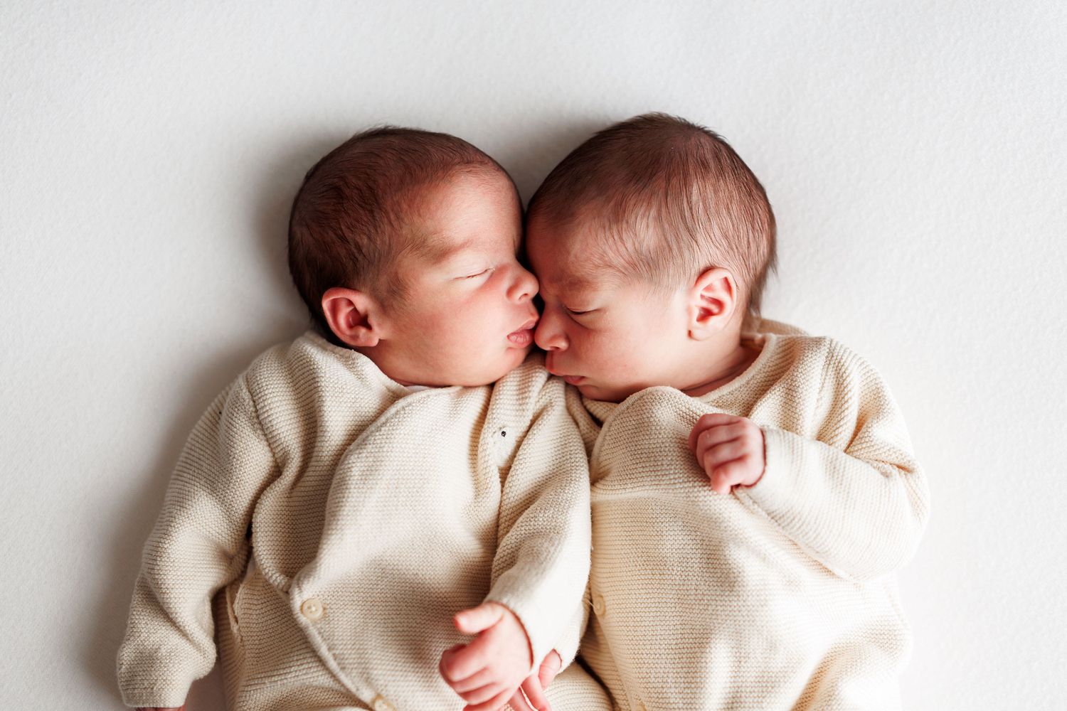 Two newborn babies in matching cream knit outfits lie side by side on a white background, their faces touching in a sweet, peaceful moment.