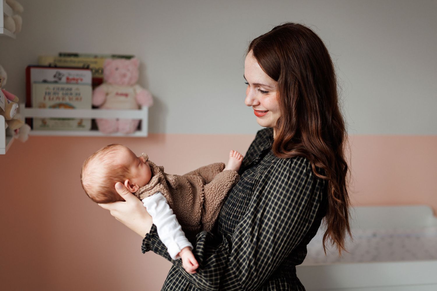 Smiling woman with long brown hair in a checkered dress gently holds a sleeping newborn baby in a cozy nursery with a pink plush toy.