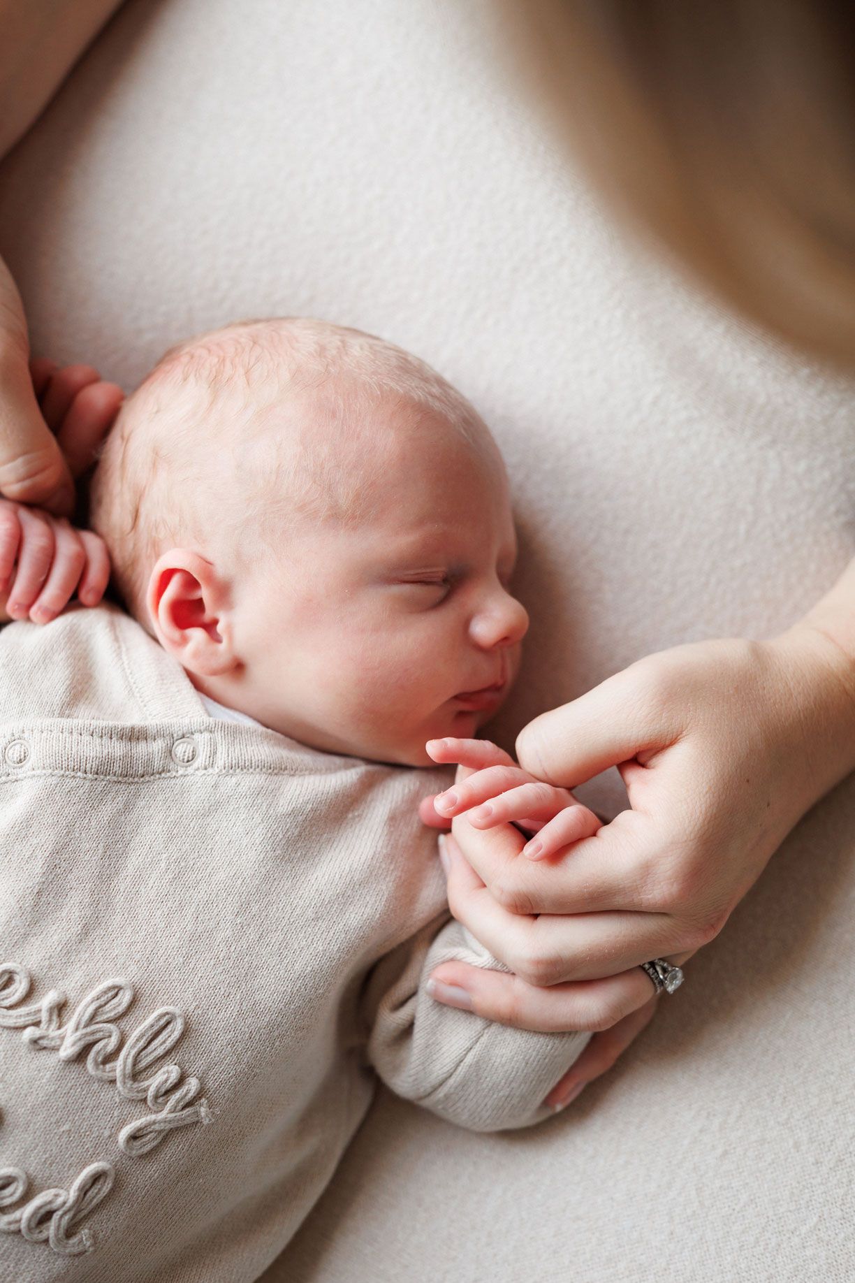 Sleeping newborn baby in a cream embroidered outfit holds a mother's finger while resting on a soft neutral blanket.