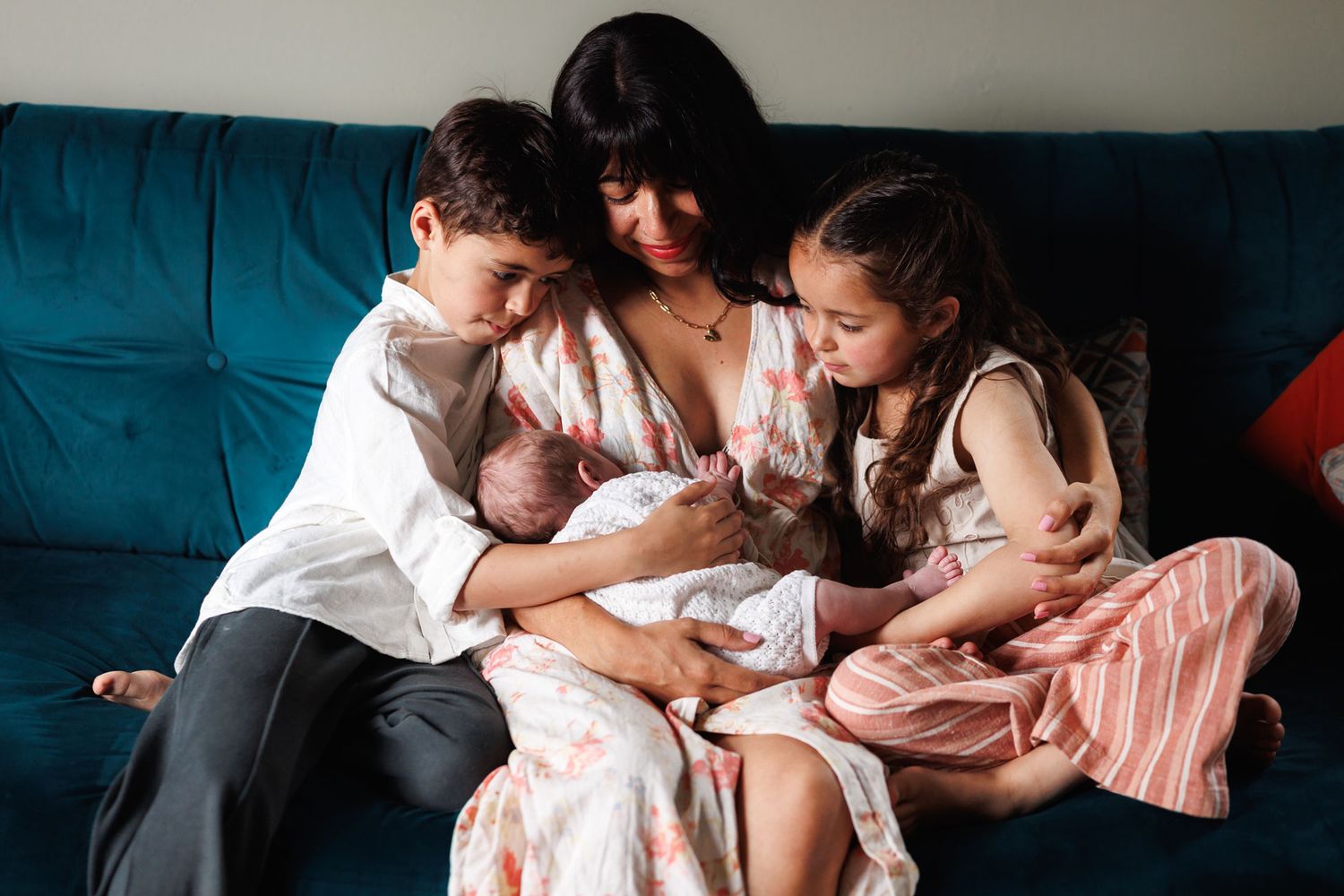 Mother sitting on teal sofa surrounded by two children admiring their newborn sibling.