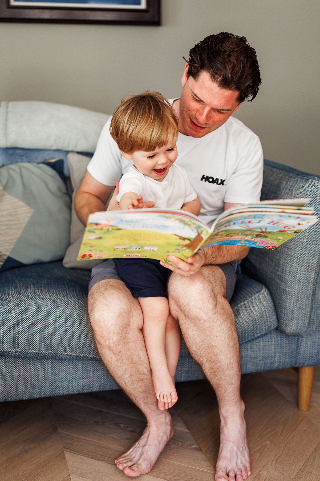 A smiling father reads a colorful picture book to his laughing toddler son while sitting on a gray sofa.