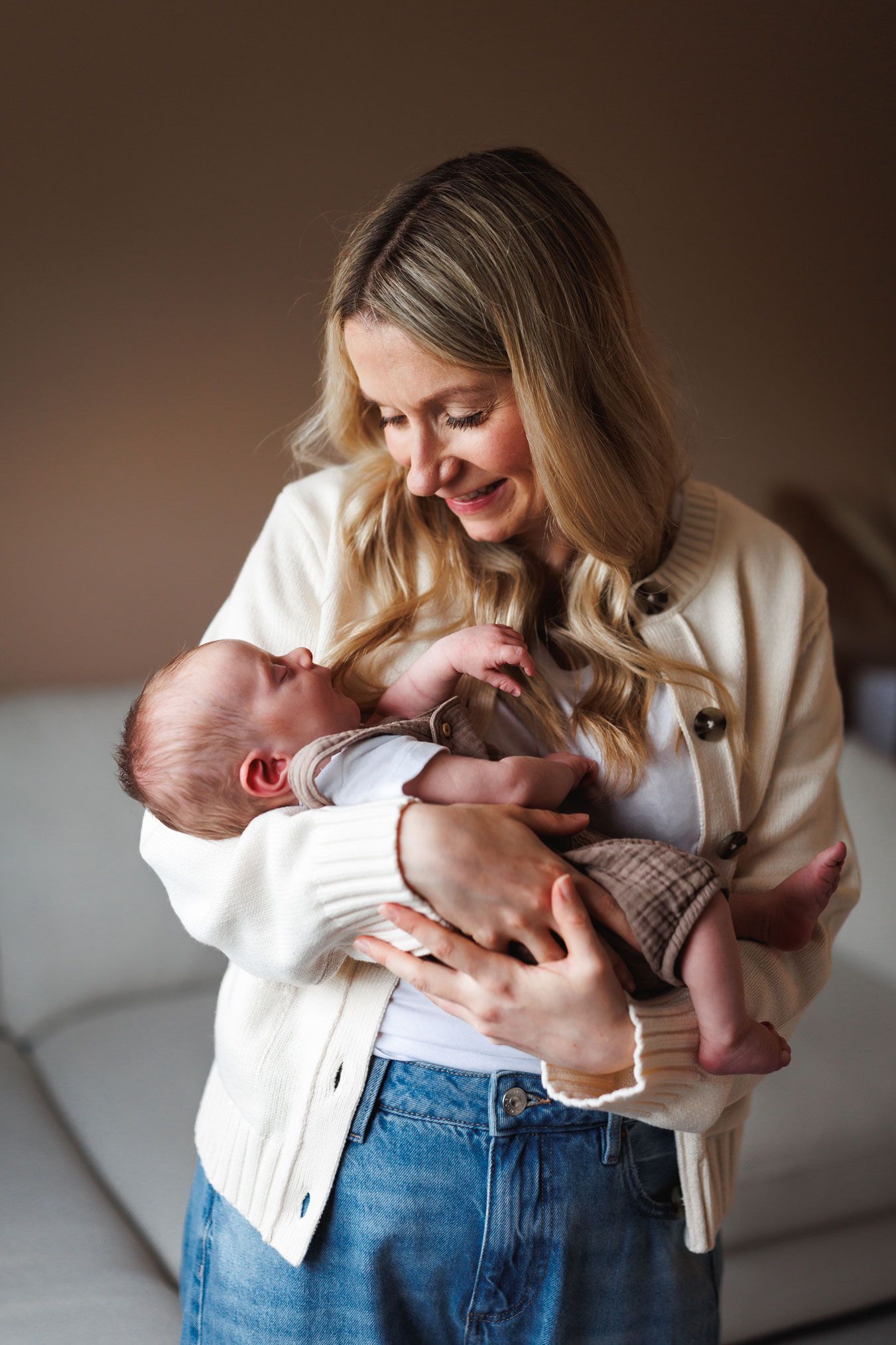 Smiling blonde mother in white cardigan and jeans tenderly holding her newborn baby at home.