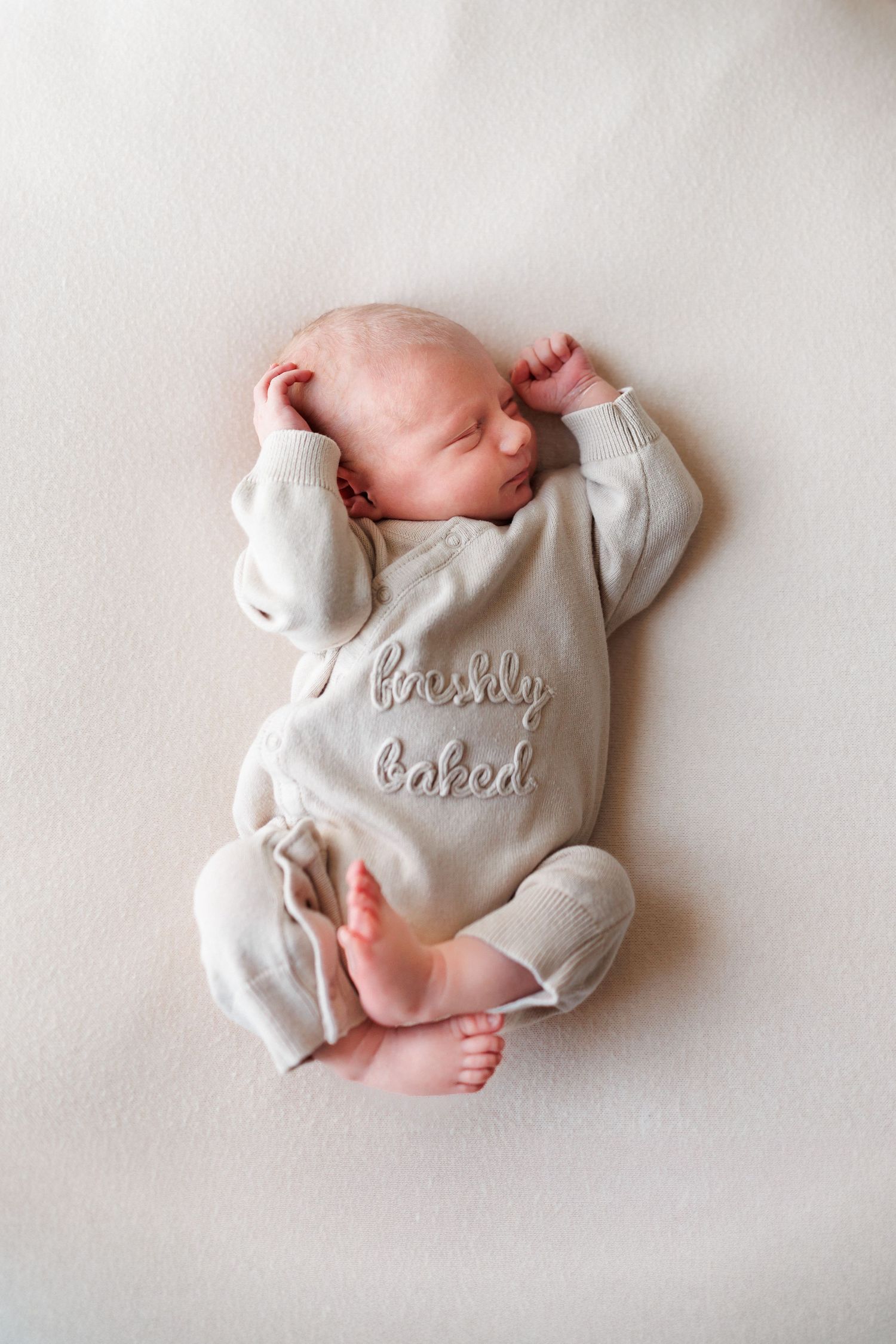 Sleeping newborn baby in a cream knit outfit embroidered with the words freshly baked, lying on a soft neutral blanket.