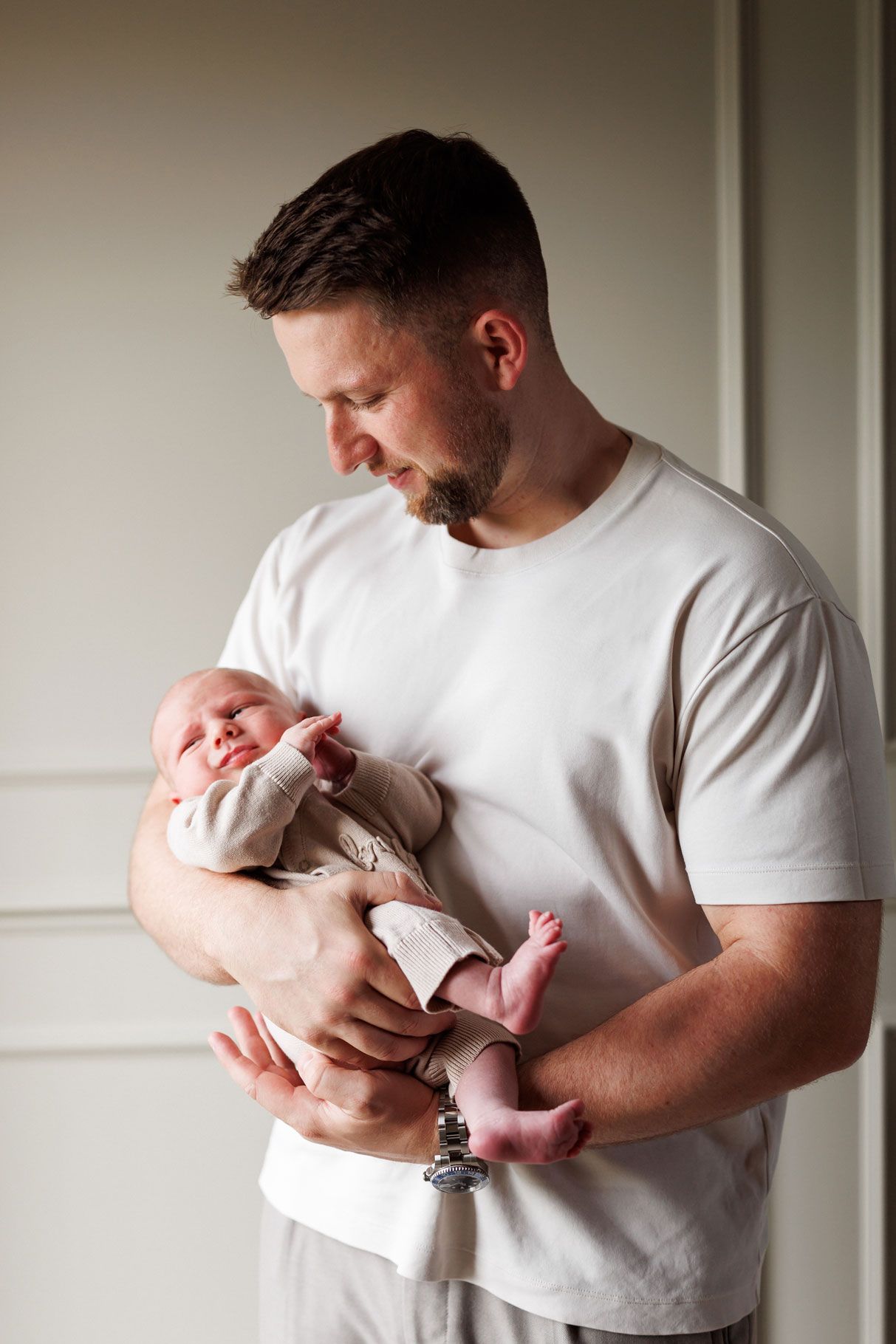 A bearded father in a white t-shirt gently cradles his sleeping newborn baby in his arms beside a bright window.