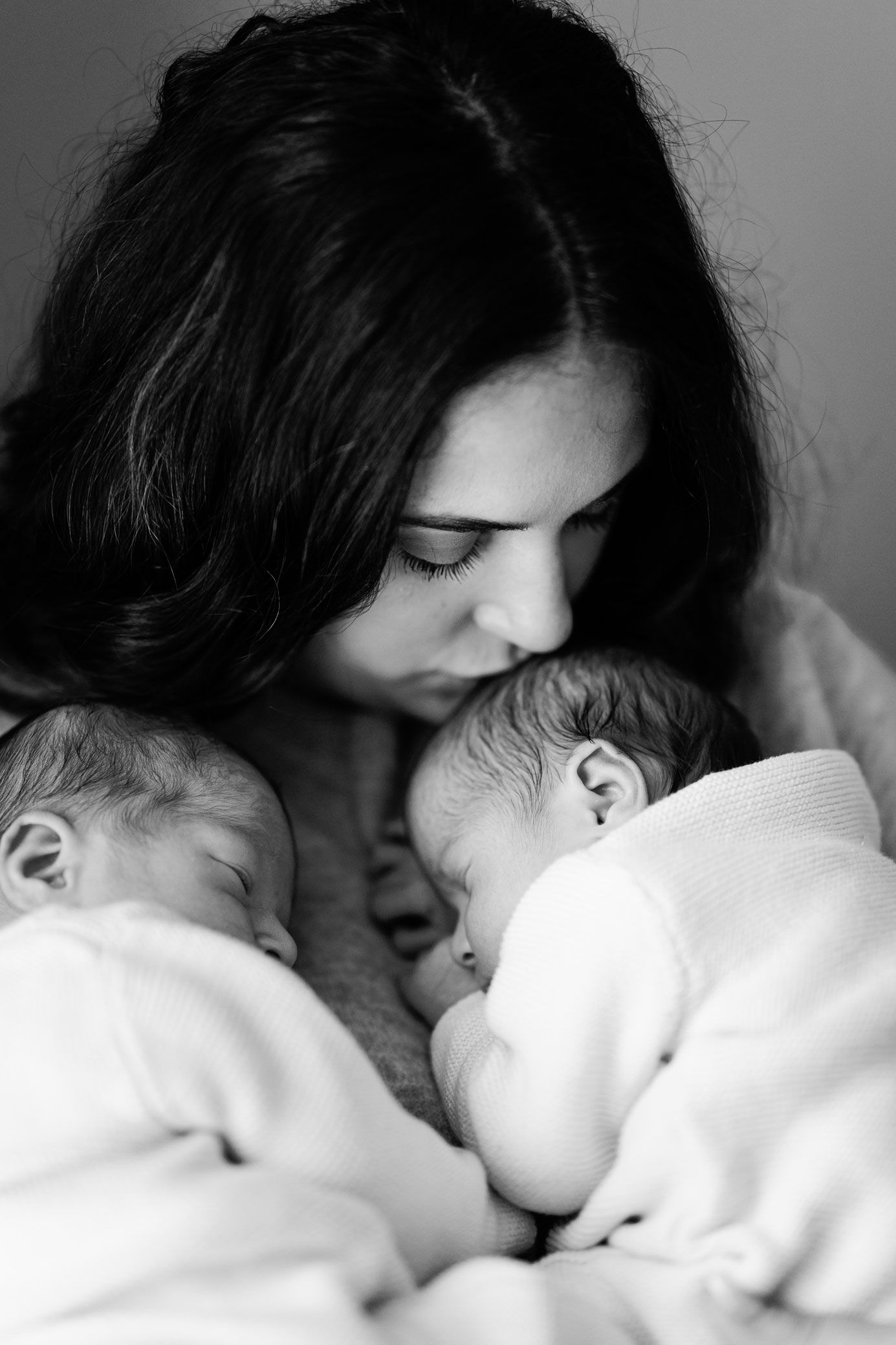 Black and white portrait of a mother with dark hair tenderly kissing the forehead of one of her two sleeping newborn twins.