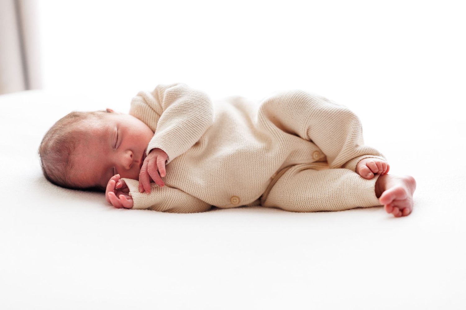 Sleeping newborn baby in a cream knit outfit curled up on a soft white background with tiny hands and feet.